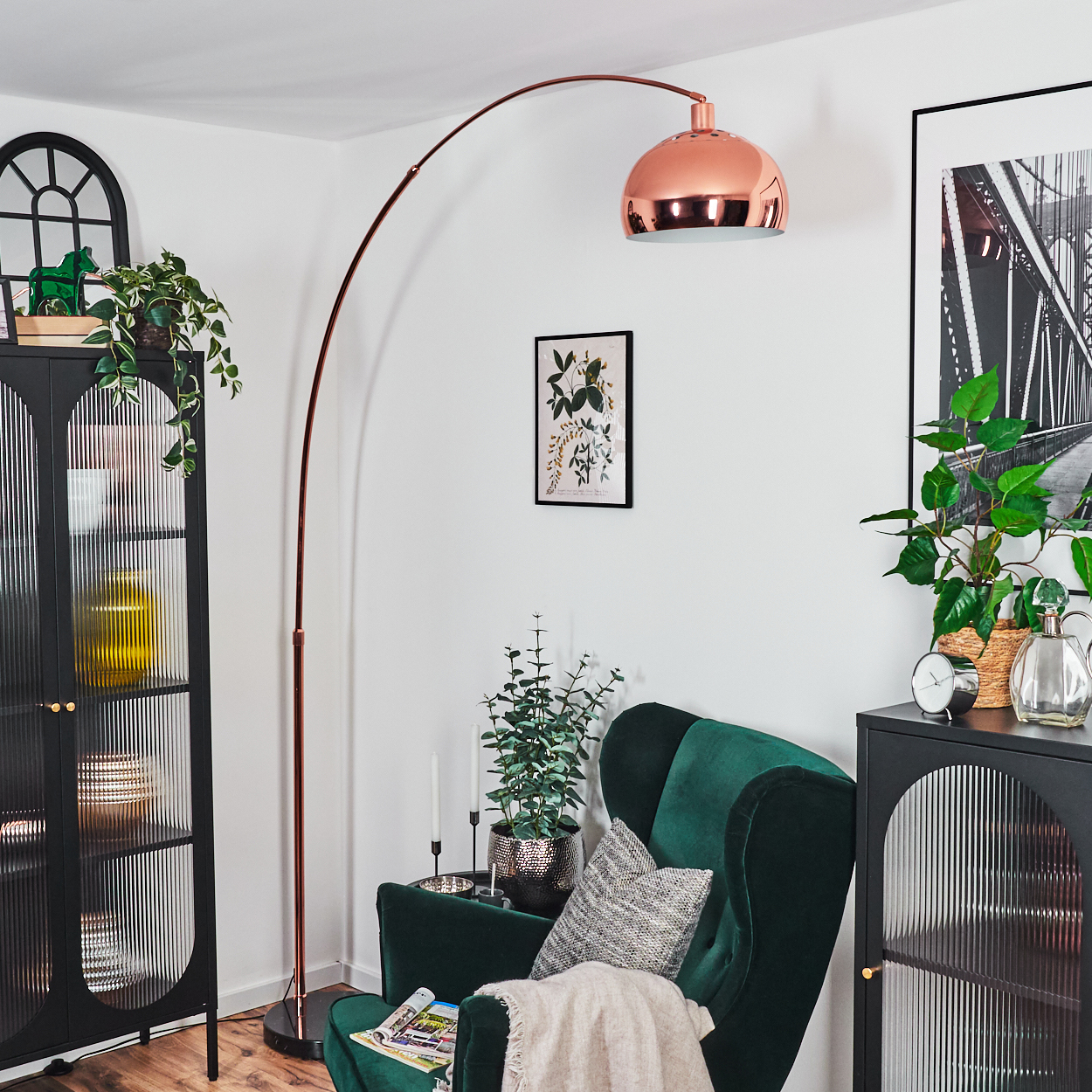 Ein Raum mit grünem Sessel, Kupferlampe und schwarzem Schrank. Pflanzen und gerahmte Bilder schmücken die weißen Wände.