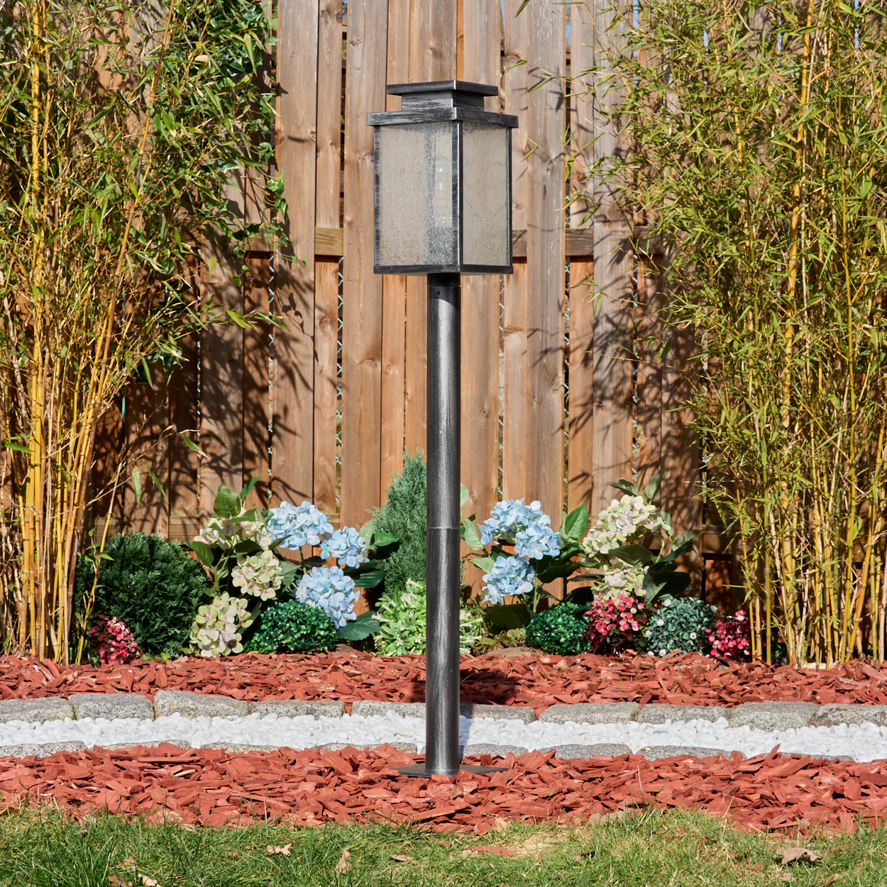 Eine Metall-Laterne steht in einem Gartenbeet mit roter Mulch und Blumen, mit einem Holzzaun und Bambus im Hintergrund.