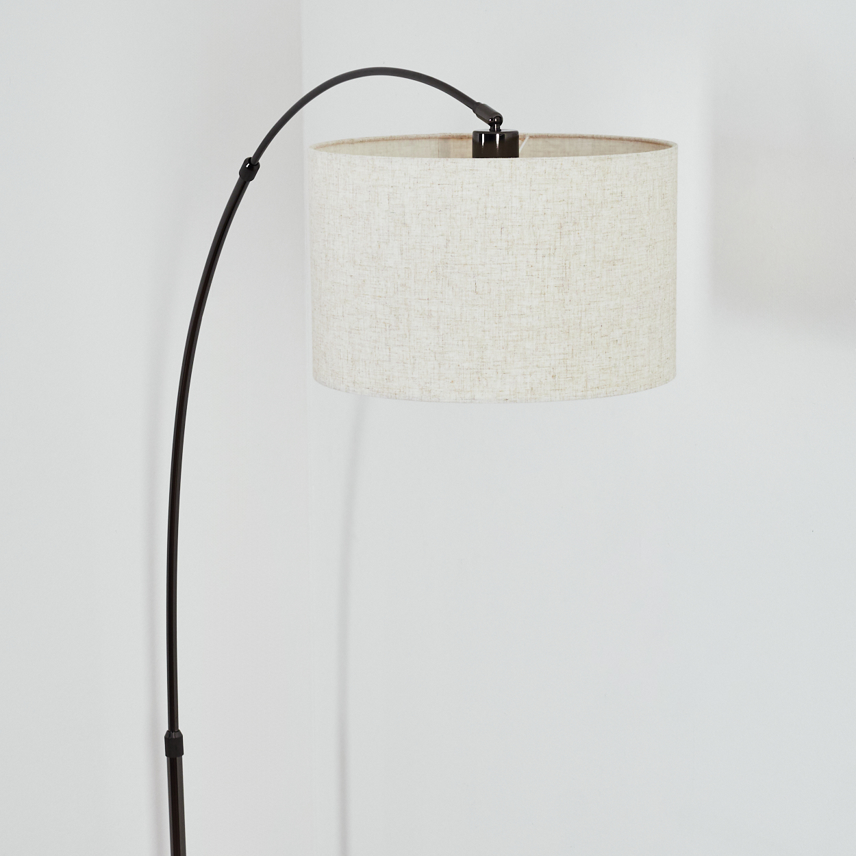Eine Stehlampe mit gebogenem schwarzem Metallarm und beigem Stoffschirm steht vor einer weißen Wand.
