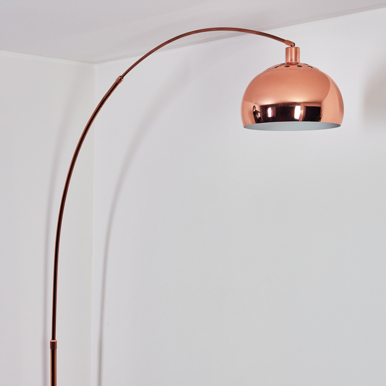 Eine kupferfarbene Bogenlampe mit gewölbtem Schirm steht vor einer weißen Wand. Der Lampenstiel ist elegant gebogen.