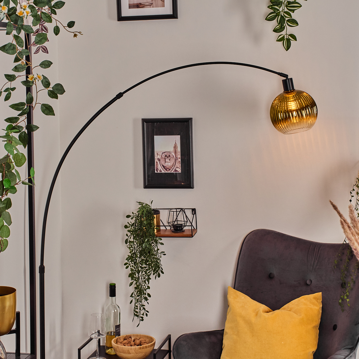 Ein Raum mit einer schwarzen Bogenlampe, einem grauen Sessel mit gelbem Kissen und einem Tisch mit Wein und Nüssen.