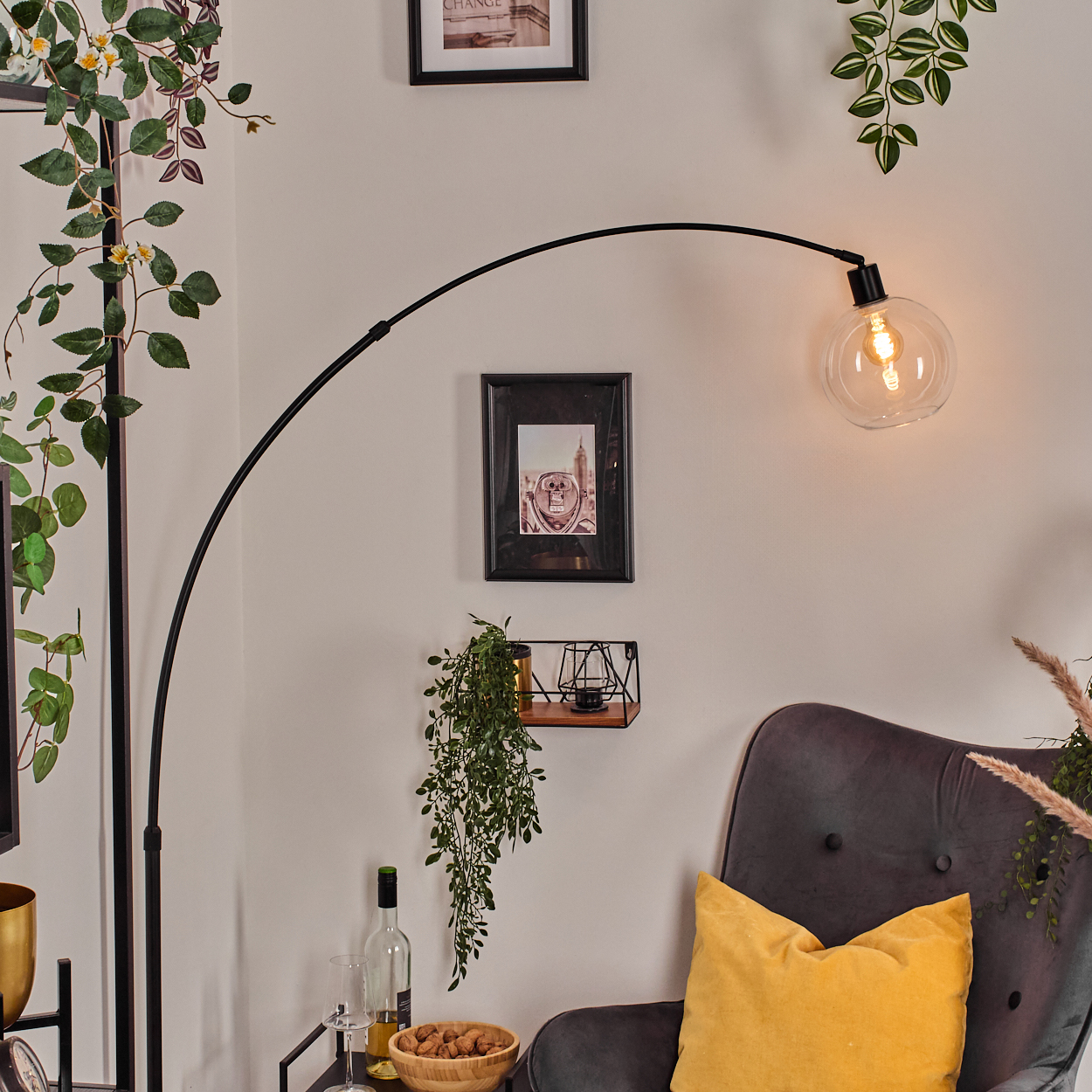 Modernes Wohnzimmer mit schwarzer Bogenlampe, grauem Sessel und gelbem Kissen. Bilder und Pflanzen schmücken die Wand.