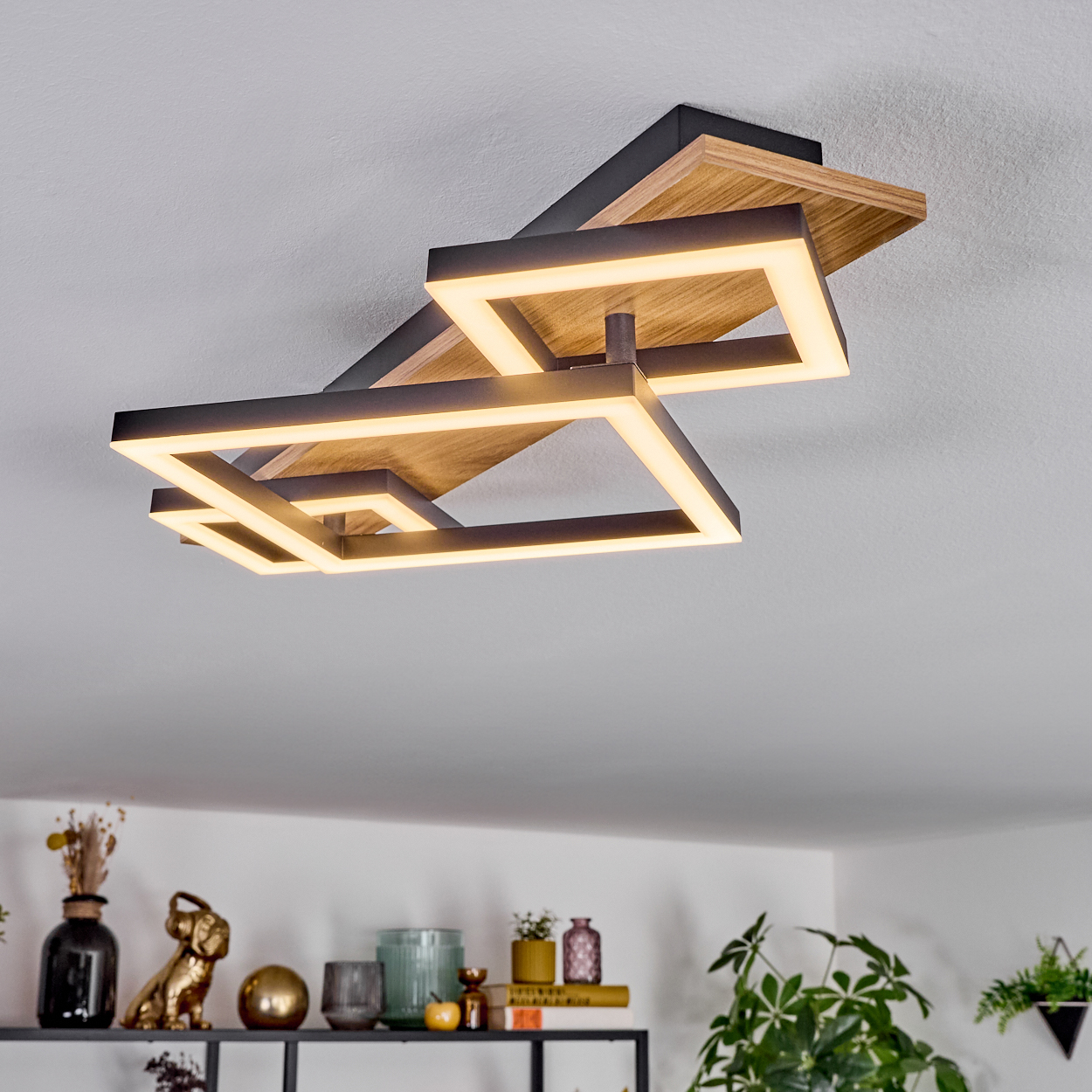 Eine moderne Deckenleuchte mit geometrischen Formen und warmer Beleuchtung hängt von der Decke. Darunter ist ein Regal mit Dekoration zu sehen.