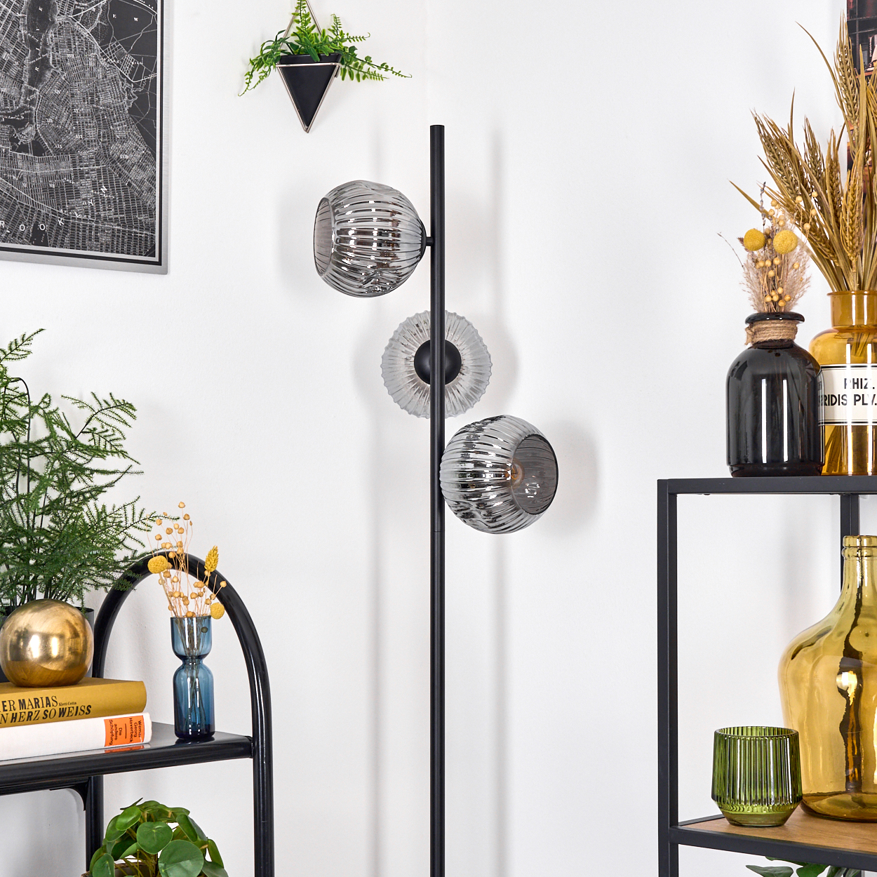 Eine Zimmerecke mit einer schwarzen Stehlampe mit drei Glasschirmen. Regale mit Vasen, Pflanzen und Büchern sind sichtbar.