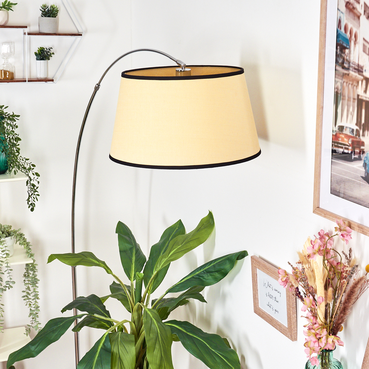 Eine Stehlampe mit beigem Lampenschirm steht neben einer großen grünen Pflanze. Ein Bilderrahmen und Blumen sind rechts.