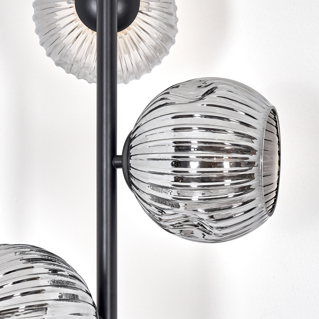 Nahaufnahme einer modernen Lampe mit drei Glasschirmen. Die Schirme sind geriffelt und silberfarben, getragen von einem schwarzen Metallrahmen. Weißer Wandhintergrund.