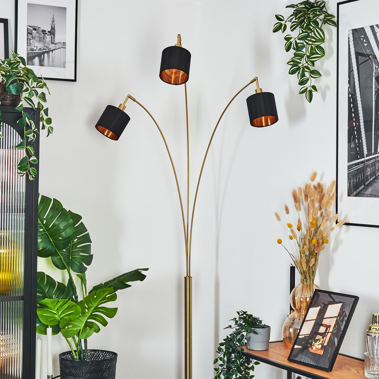 Goldene Stehlampe mit drei schwarzen Schirmen. Pflanzen und gerahmte Bilder schmücken einen Raum mit weißen Wänden. Ein dekoratives Regal ist sichtbar.