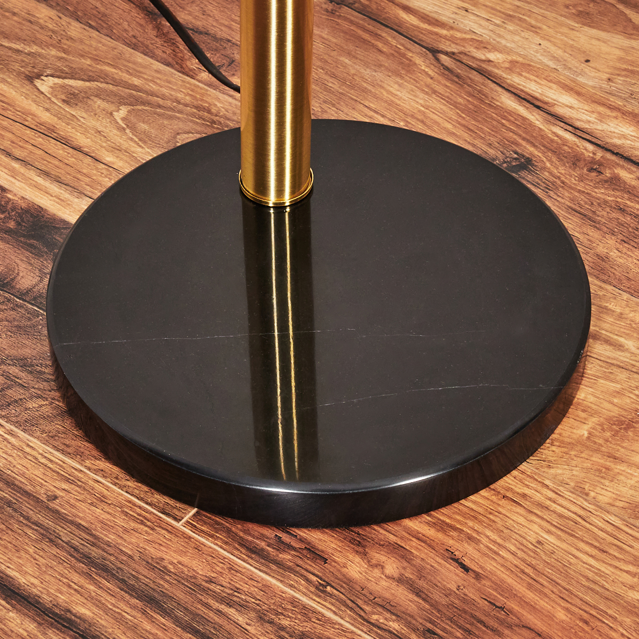 Nahaufnahme eines Lampenfußes. Der Sockel ist eine schwarze, kreisförmige Marmorplatte, die einen goldenen Metallpfosten trägt. Der Boden ist aus Holz.