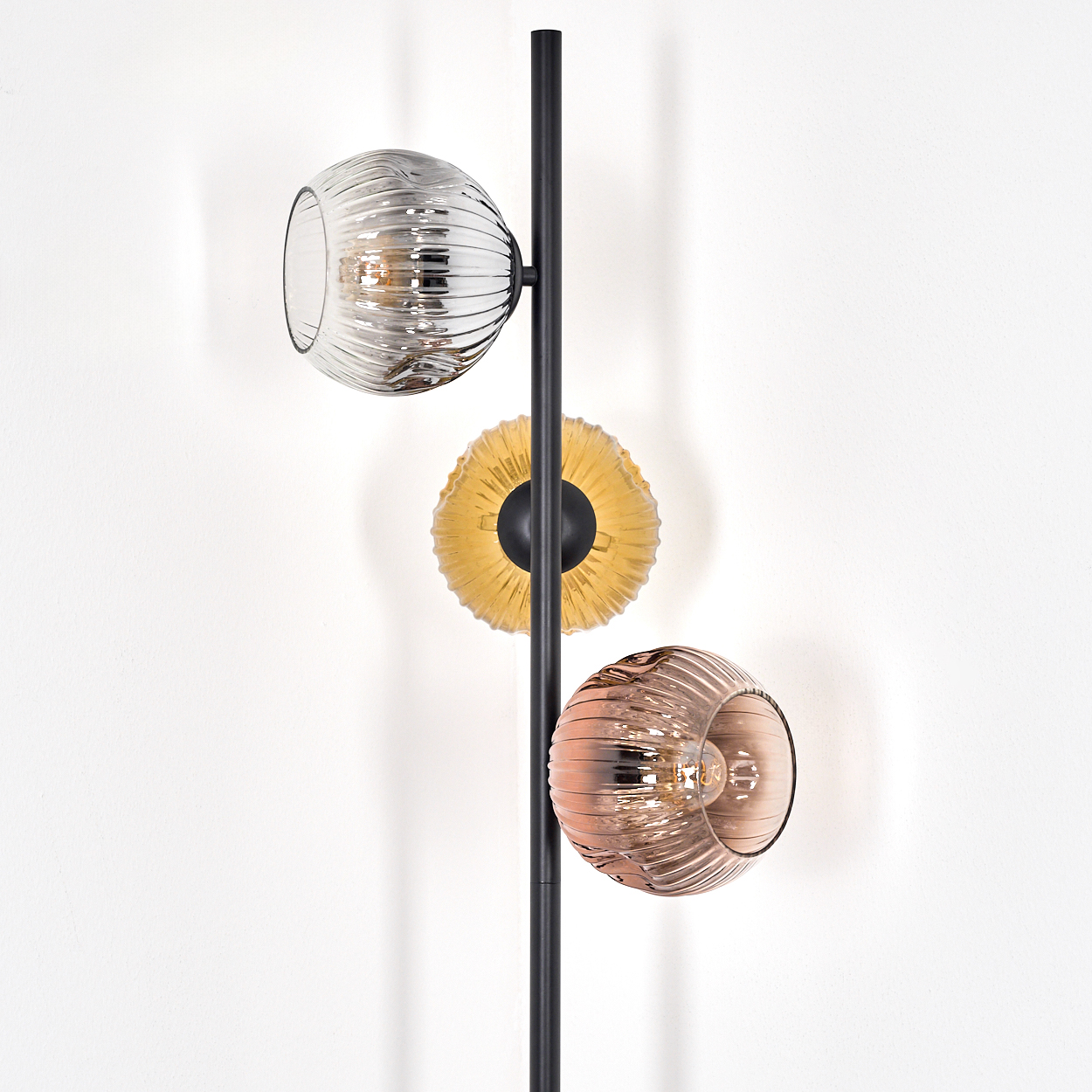 Eine moderne Lampe mit drei Glasschirmen in verschiedenen Farben und Formen, an einer schwarzen Metallstange, vor einer weißen Wand.