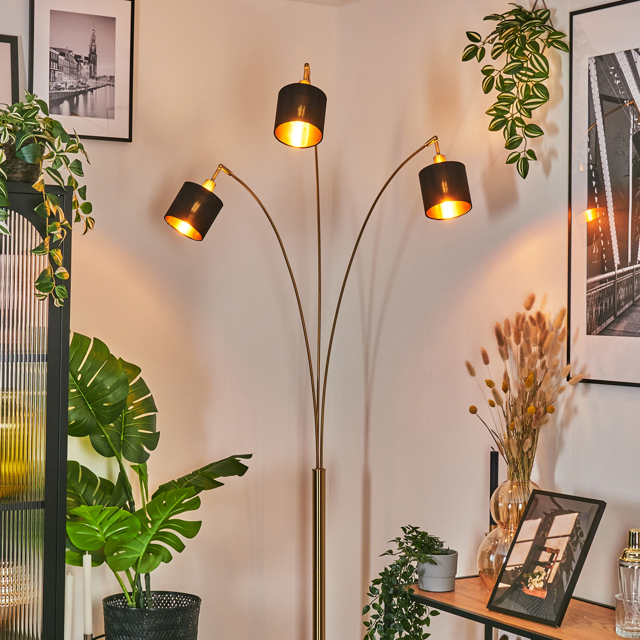 Eine goldene Stehlampe mit drei schwarzen Schirmen beleuchtet einen Raum mit Pflanzen, Bildern und einem Holzregal.