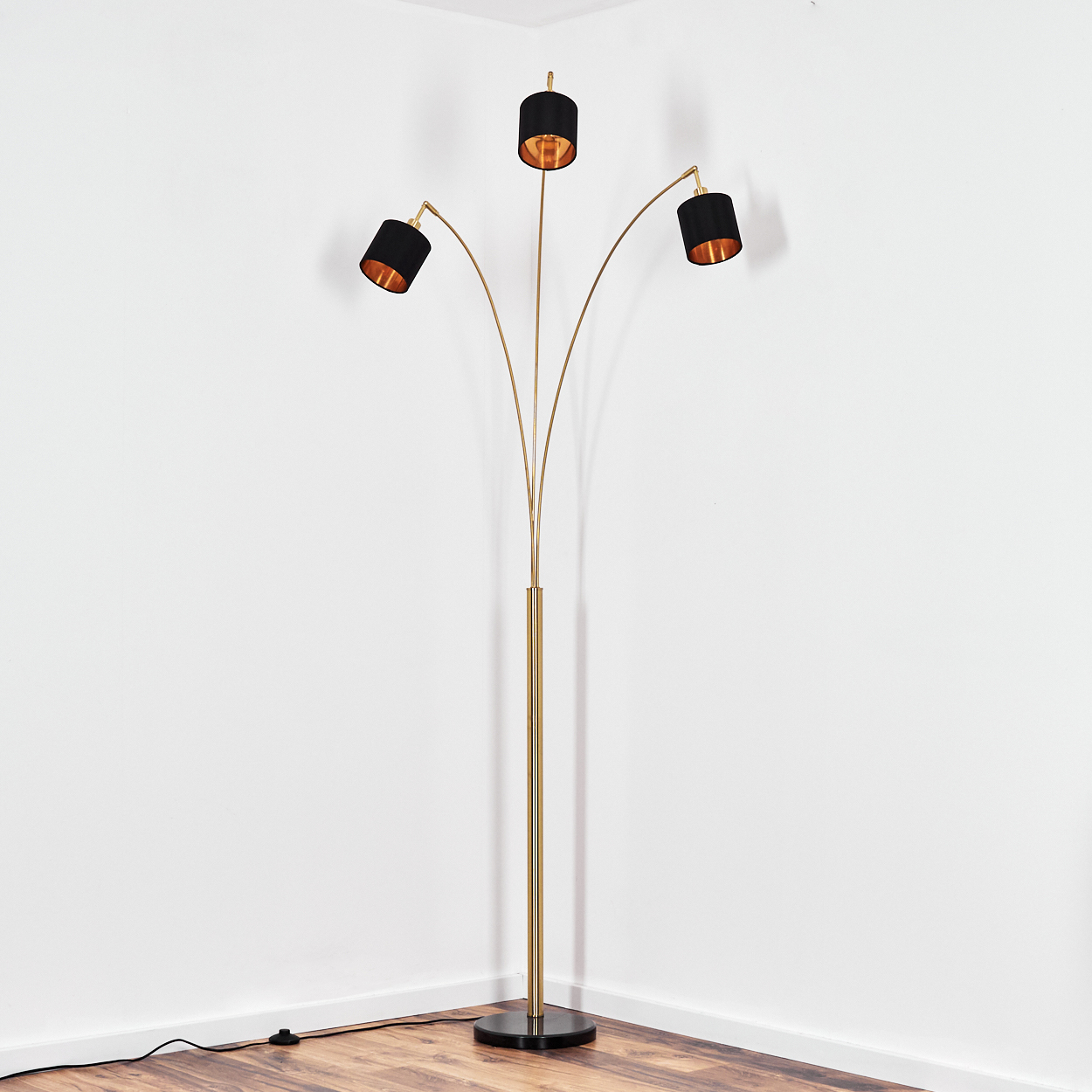 Eine goldene Stehlampe mit drei schwarzen Schirmen. Sie steht auf einem Holzboden neben einer weißen Wand.