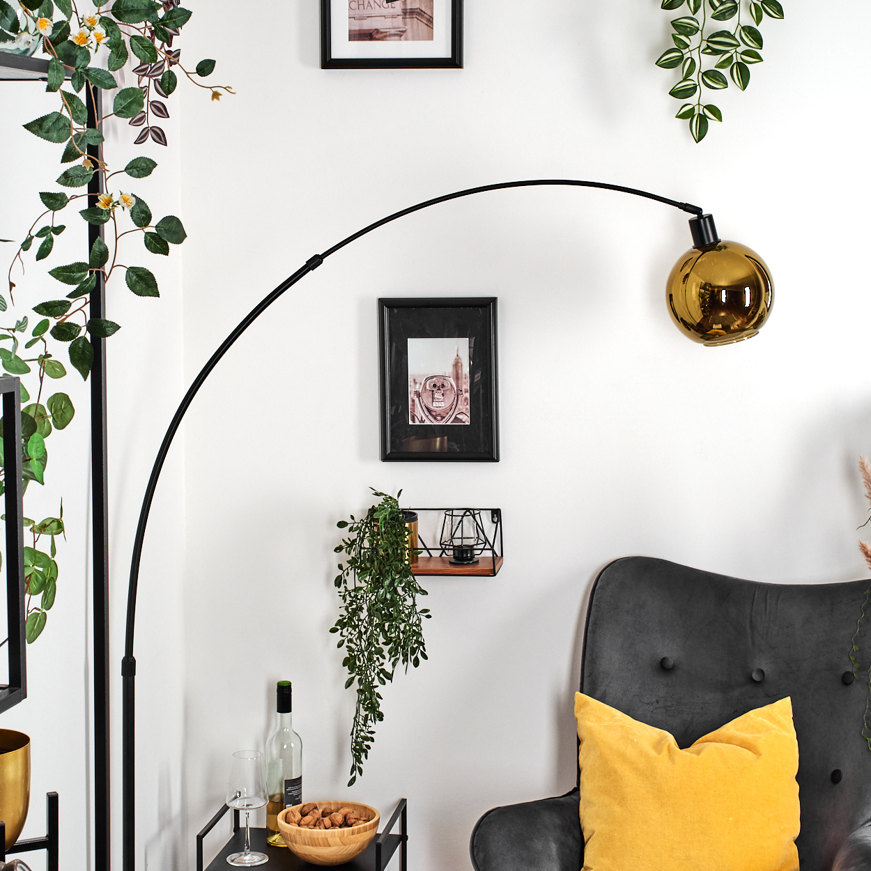 Ein Raum mit einer schwarzen Bogenlampe, Pflanzen, einem schwarzen Bild mit Rahmen, einem grauen Sessel und einem gelben Kissen.