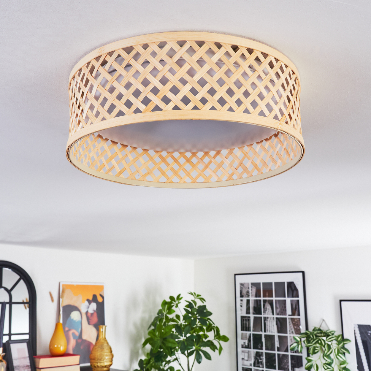 Eine Deckenlampe aus geflochtenem Bambus hängt in einem Raum mit Kunst und Pflanzen. Die Lampe ist kreisförmig mit Gitterdesign.