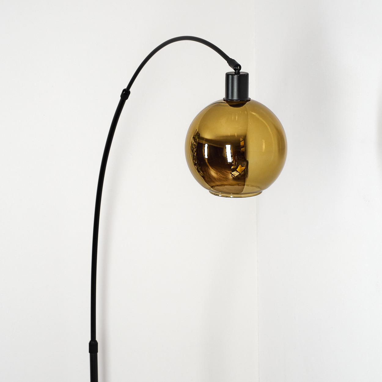 Eine moderne Stehlampe mit einem schwarzen, gebogenen Arm und einem goldgelben, kugelförmigen Lampenschirm vor einer weißen Wand.