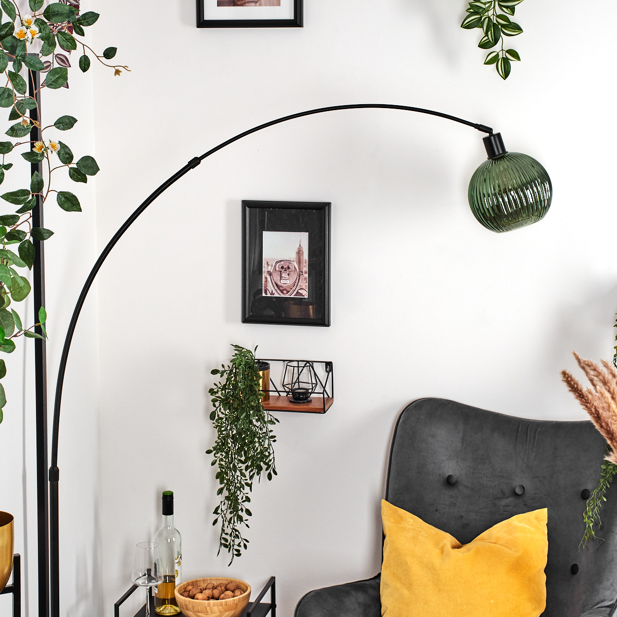 Ein Raum mit einer schwarzen Bogenlampe, einem grauen Sessel mit gelbem Kissen und Pflanzen. Ein Bild hängt an der weißen Wand.