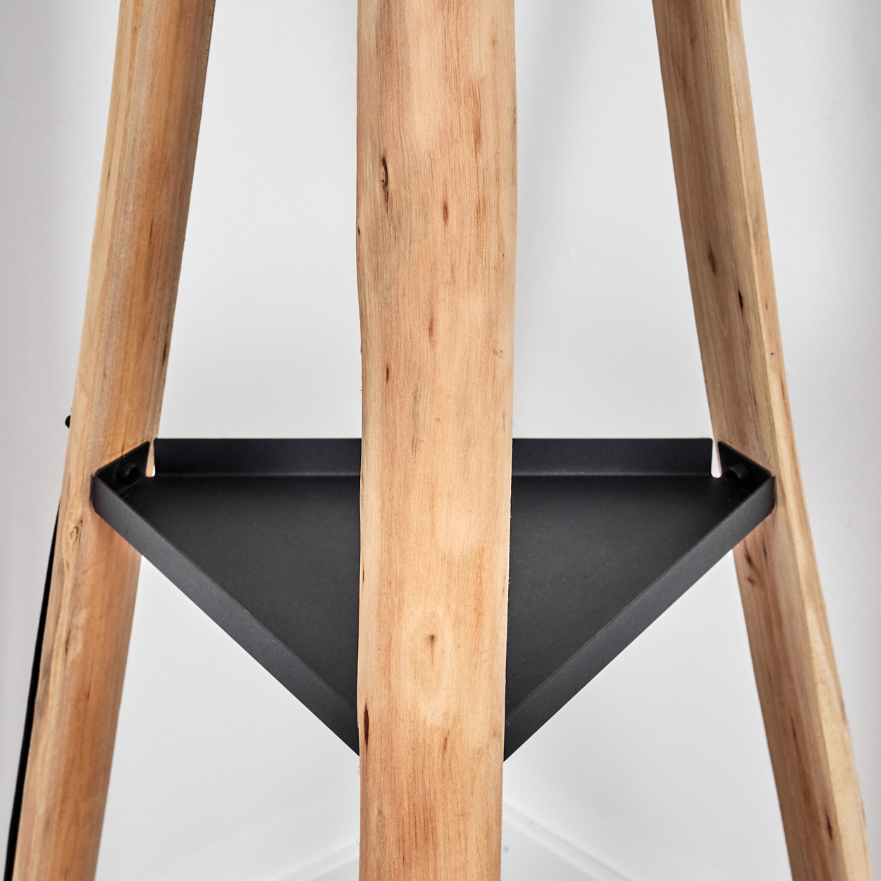 Eine Holz-Dreibein-Lampe mit einem schwarzen dreieckigen Regal. Das Holz hat eine natürliche Maserung. Der Hintergrund ist eine weiße Wand.
