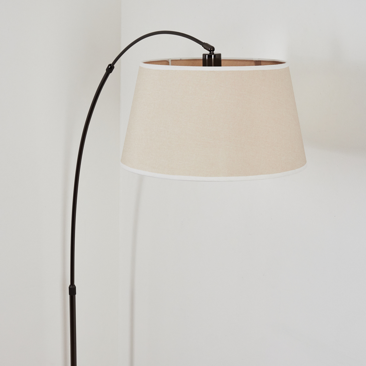 Ein beigefarbener Lampenschirm mit weißem Rand hängt an einem schwarzen, gebogenen Lampenarm. Die Lampe steht vor einer weißen Wand.