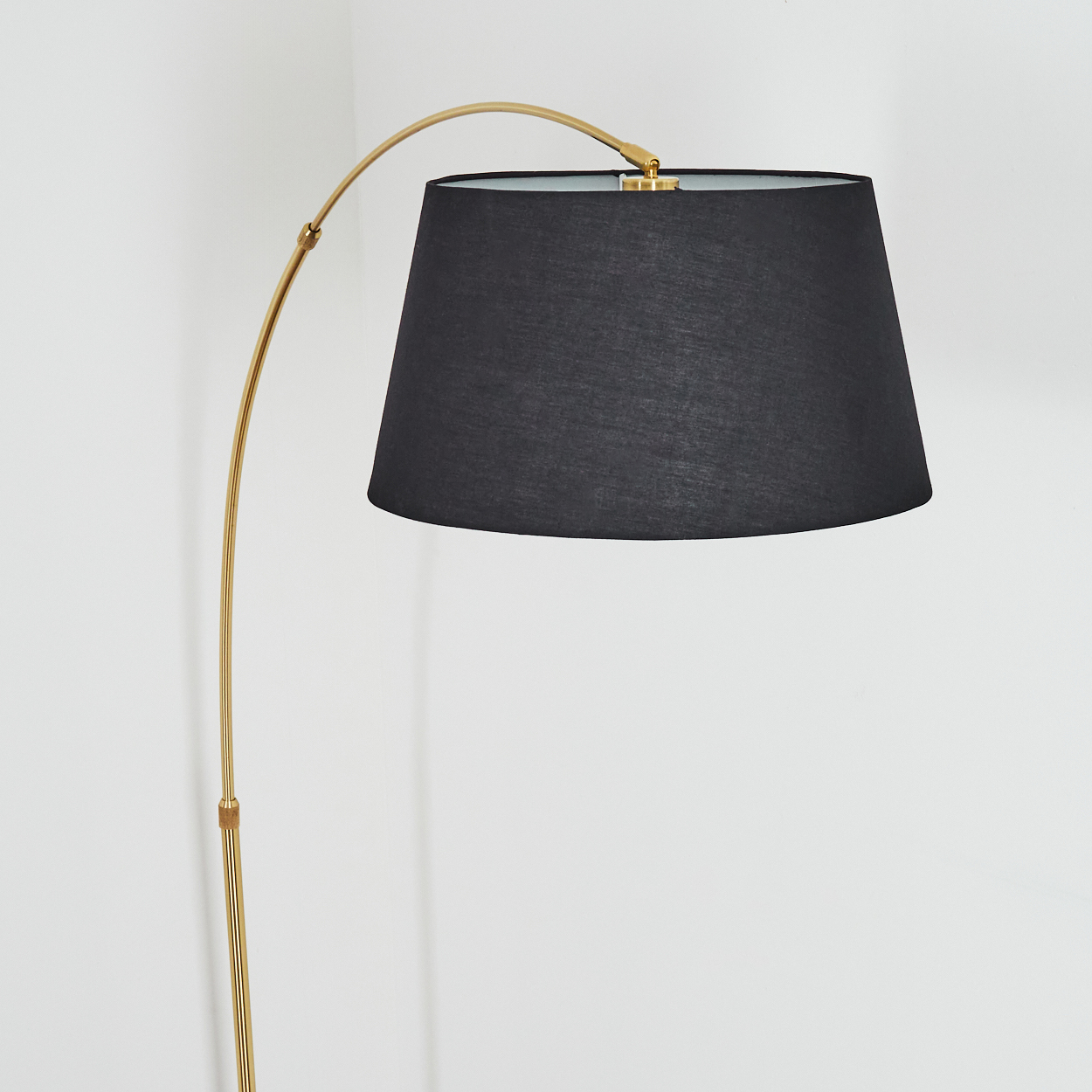 Eine goldene Stehlampe mit schwarzem Lampenschirm steht in der Ecke eines weißen Raumes. Die Lampe hat einen gebogenen Arm.