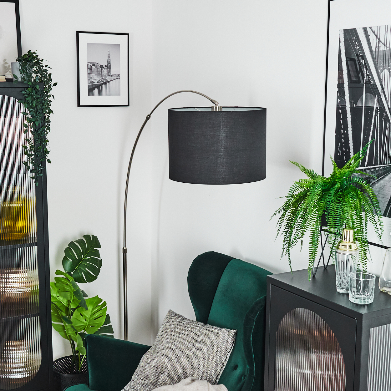 Eine Zimmerecke mit grünem Stuhl, Stehlampe und schwarzem Schrank mit Pflanzen und Bildern an der Wand.