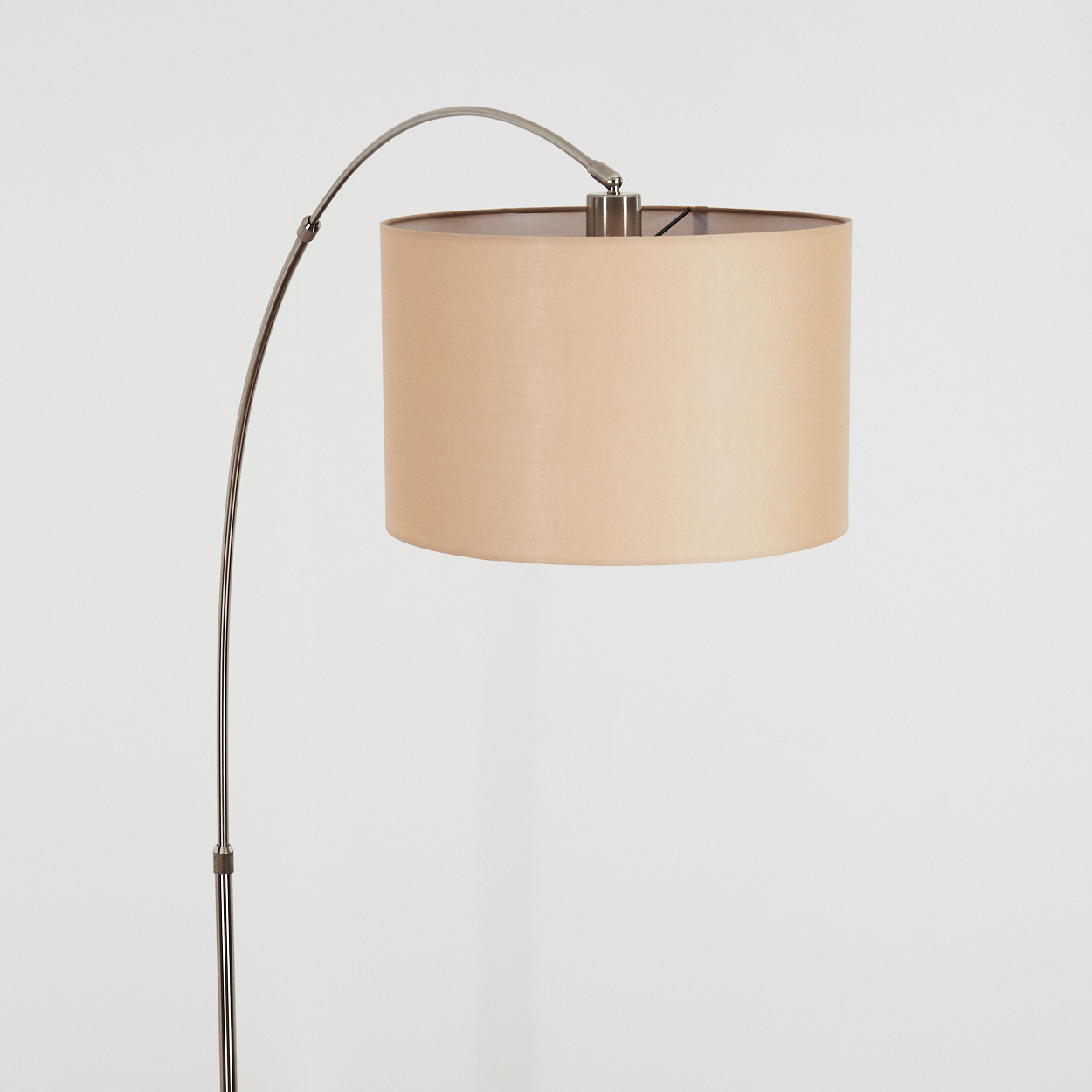 Moderne Bogenstehlampe mit beigem Lampenschirm. Die Lampe hat ein silbernes Metallgestell, der Hintergrund ist eine weiße Wand.