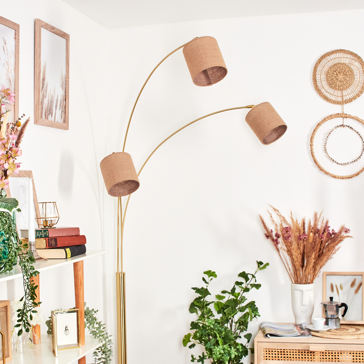 Ein Raum mit einer goldenen Stehlampe mit drei Lampenschirmen. Es gibt ein weißes Regal mit Büchern und Pflanzen und einen Schrank mit Blumen.