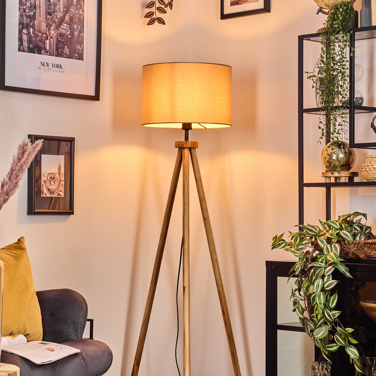 Eine beleuchtete Stehlampe aus Holz mit beigem Schirm steht in einem Raum. Im Hintergrund sind Bilder und Pflanzen. Links ist ein Sofa mit gelbem Kissen.