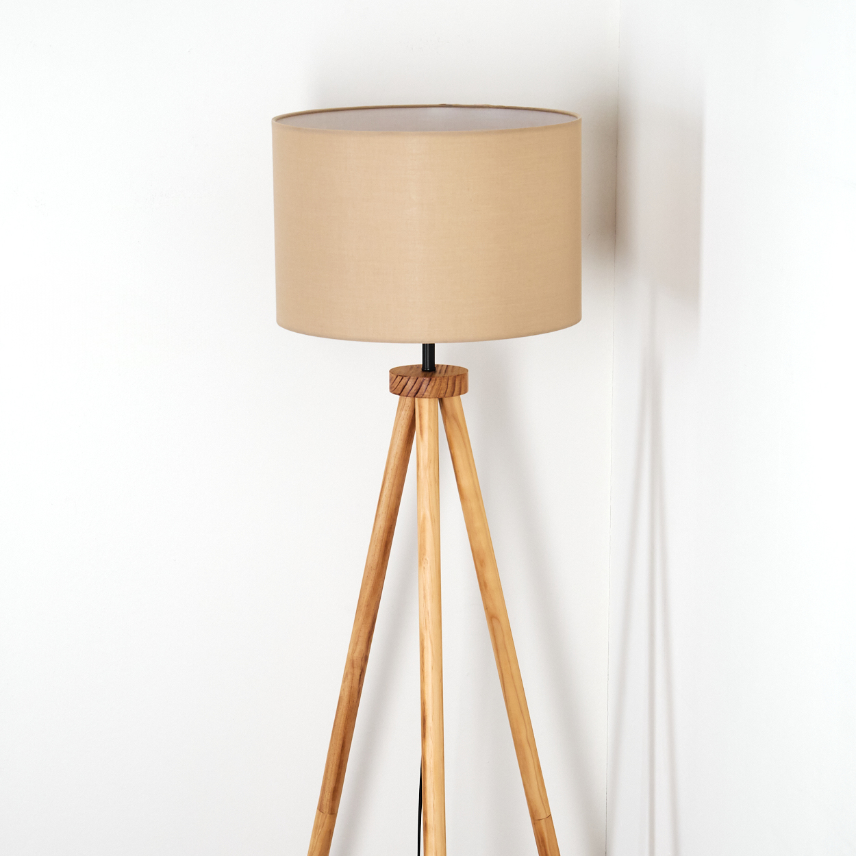 Ein beigefarbener Lampenschirm sitzt auf einem hölzernen Stativ-Stehlampenfuß. Der Hintergrund ist eine weiße Wand und der Schatten der Lampe.