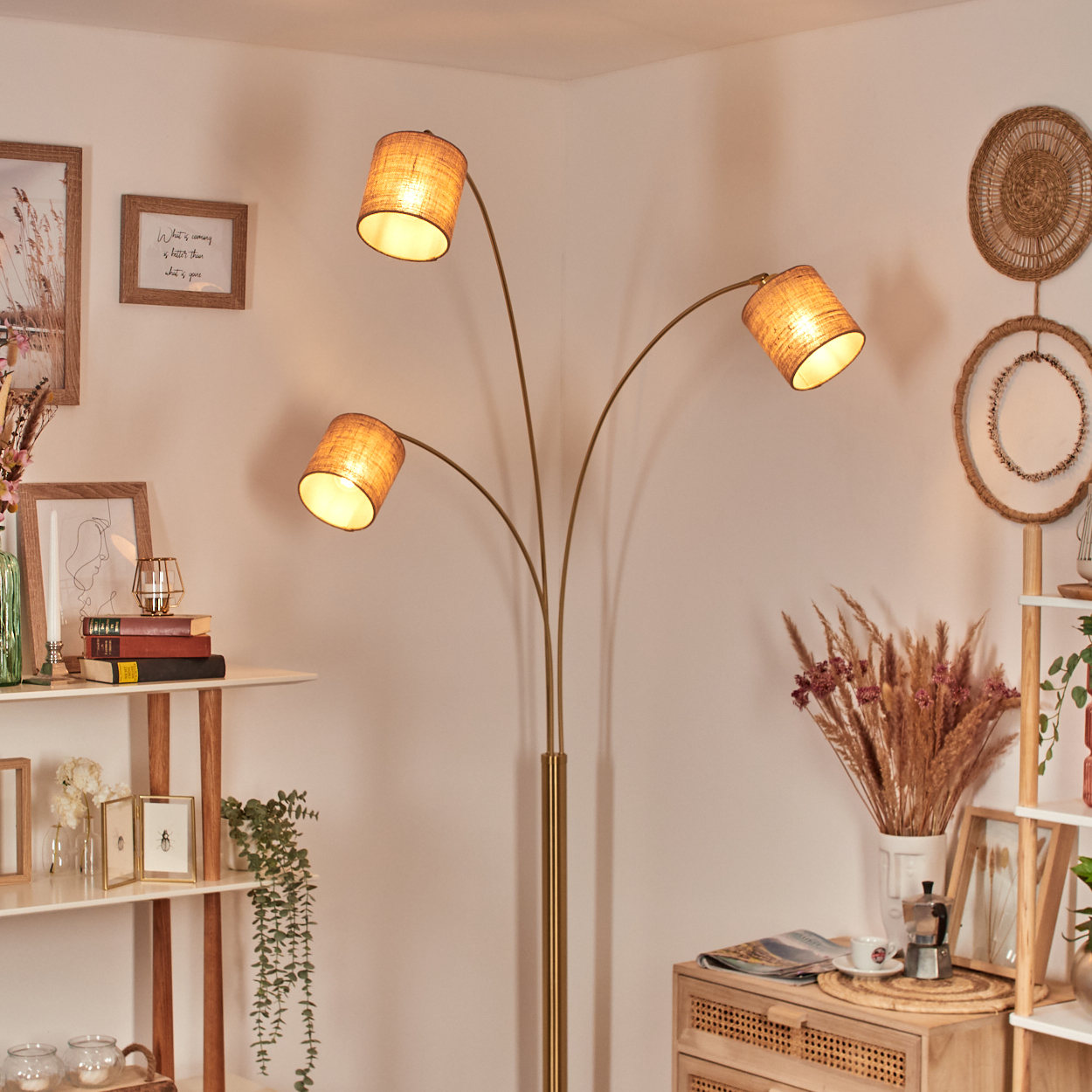 Eine Stehlampe mit drei Lampenschirmen in einem Raum. Regale mit Dekorationen, Trockenblumen und ein Holzschrank sind ebenfalls zu sehen.