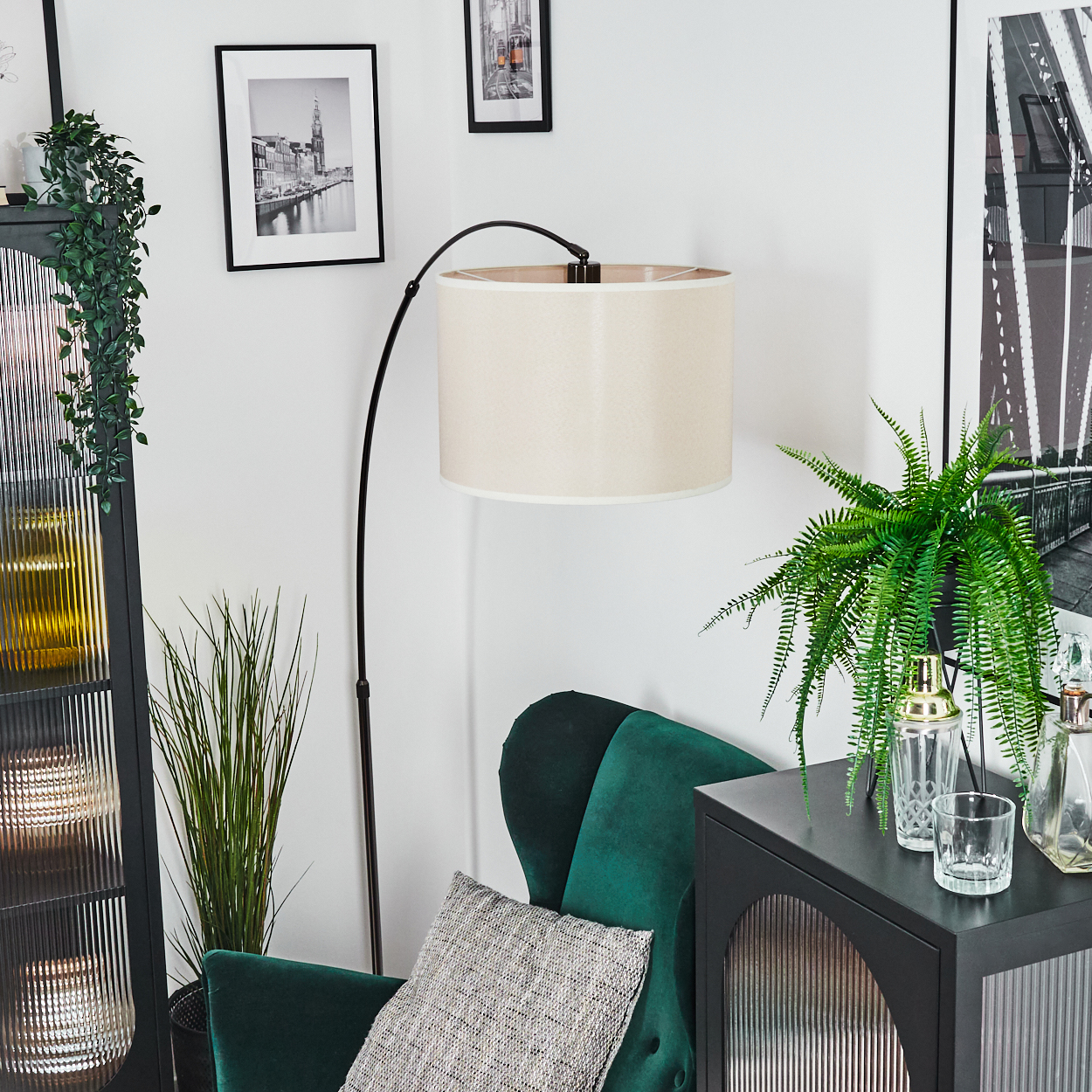 Wohnzimmer mit grünem Sessel, Lampe, Bildern und Pflanzen. Ein schwarzer Schrank hat einen Farn, einen Cocktailshaker und Gläser.