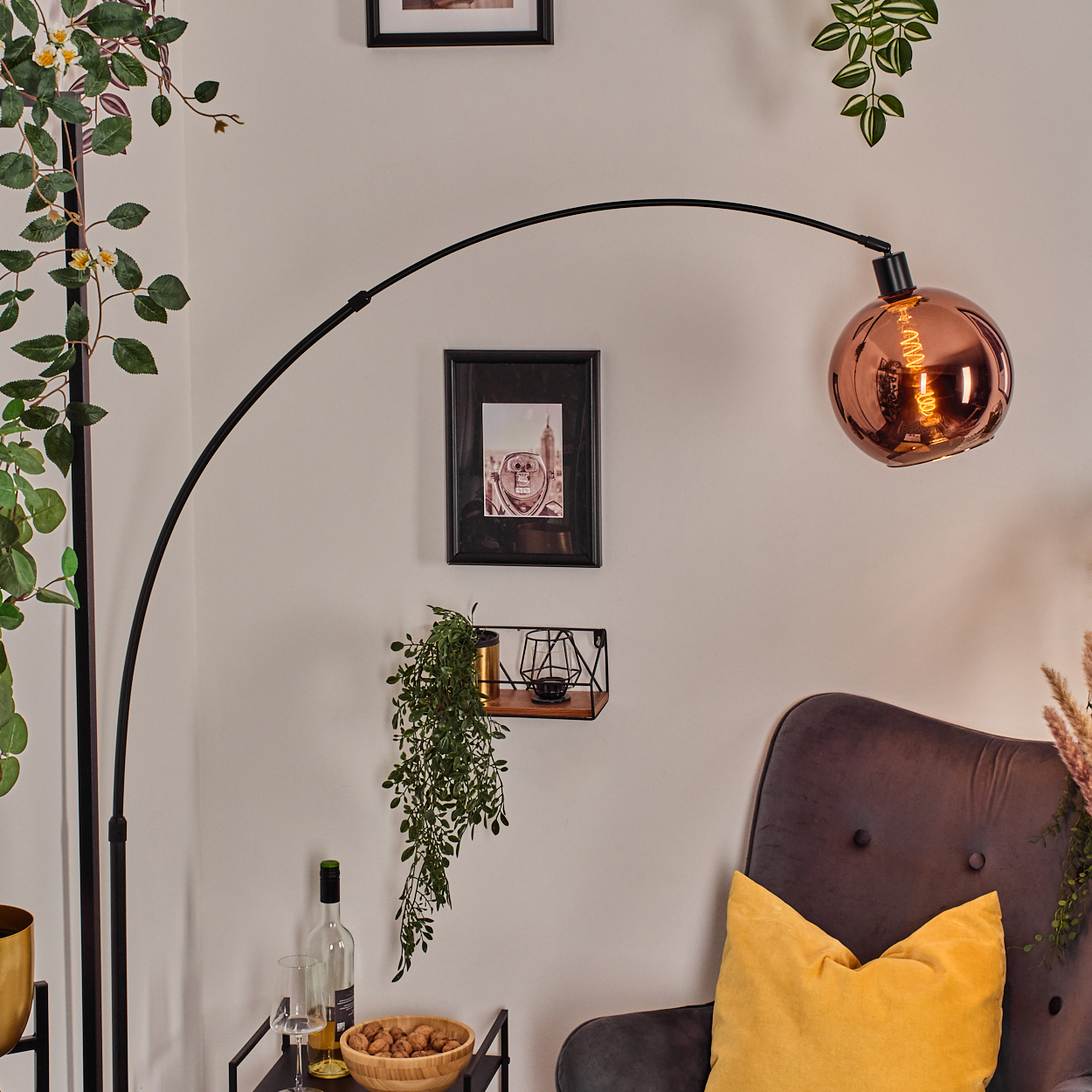 Ein Raum mit einer schwarzen Bogenlampe, einem kupferfarbenen Schirm und einem dunkelgrauen Sessel mit gelbem Kissen. Pflanzen und eine Weinflasche sind vorhanden.