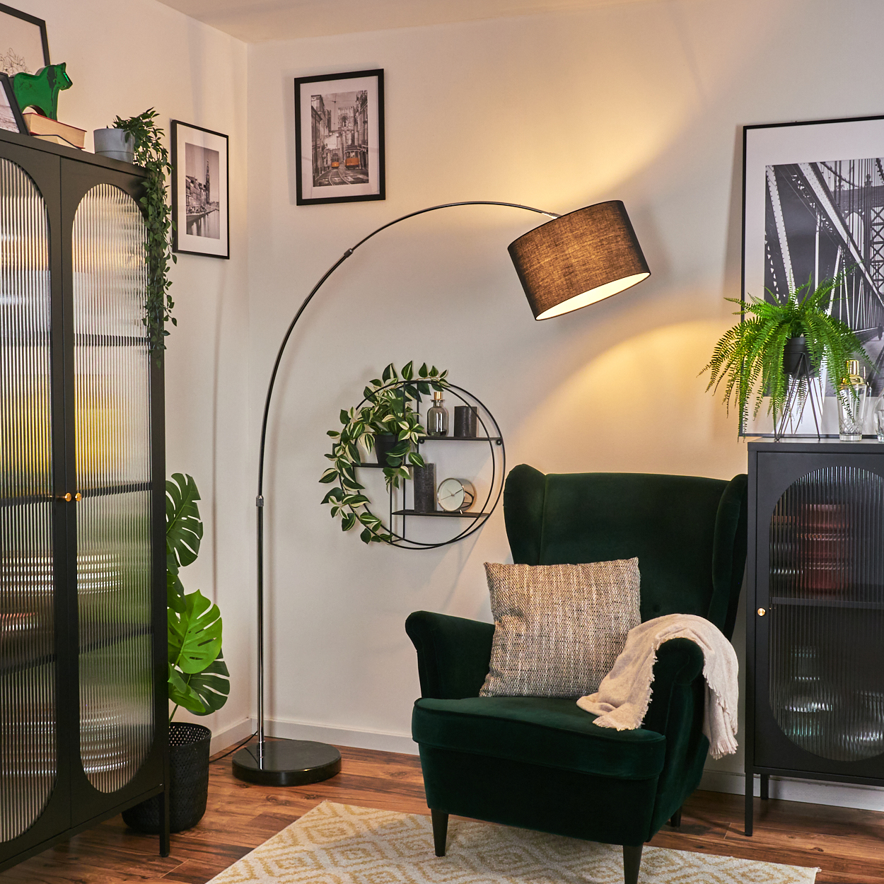 Ein grüner Samt-Sessel mit einem Wurfkissen und einer Decke steht unter einer Lampe. Schwarze Schränke und Wandkunst sind ebenfalls zu sehen.