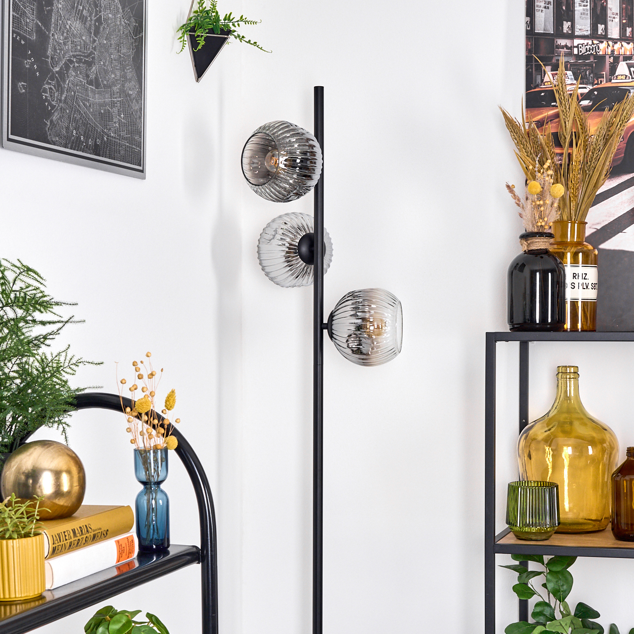 Ein modernes Zimmer mit einer schwarzen Stehlampe mit drei Glasschirmen. Auf Regalen stehen Vasen, Bücher und Pflanzen vor einer weißen Wand.