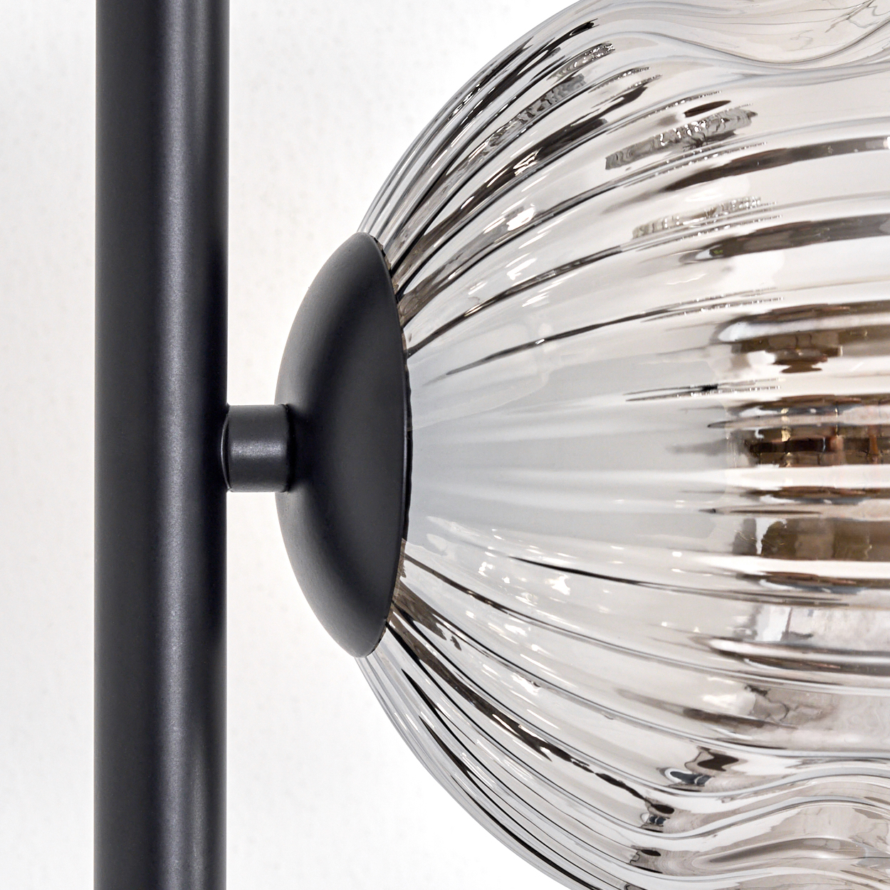 Nahaufnahme einer modernen Lampe. Ein schwarzer Metallpfosten trägt eine Glaskugel mit vertikalen Rippen. Der Hintergrund ist weiß.