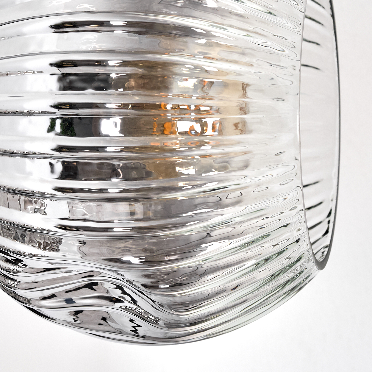 Nahaufnahme eines transparenten Glasobjekts mit welliger, geriffelter Textur. Das Glas reflektiert Licht, mit weißem Hintergrund.