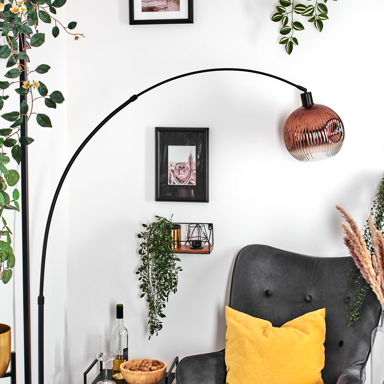 Modernes Wohnzimmer mit schwarzer Bogenlampe, grauem Sessel, gelbem Kissen und dekorativen Pflanzen. Bilder hängen an der weißen Wand.