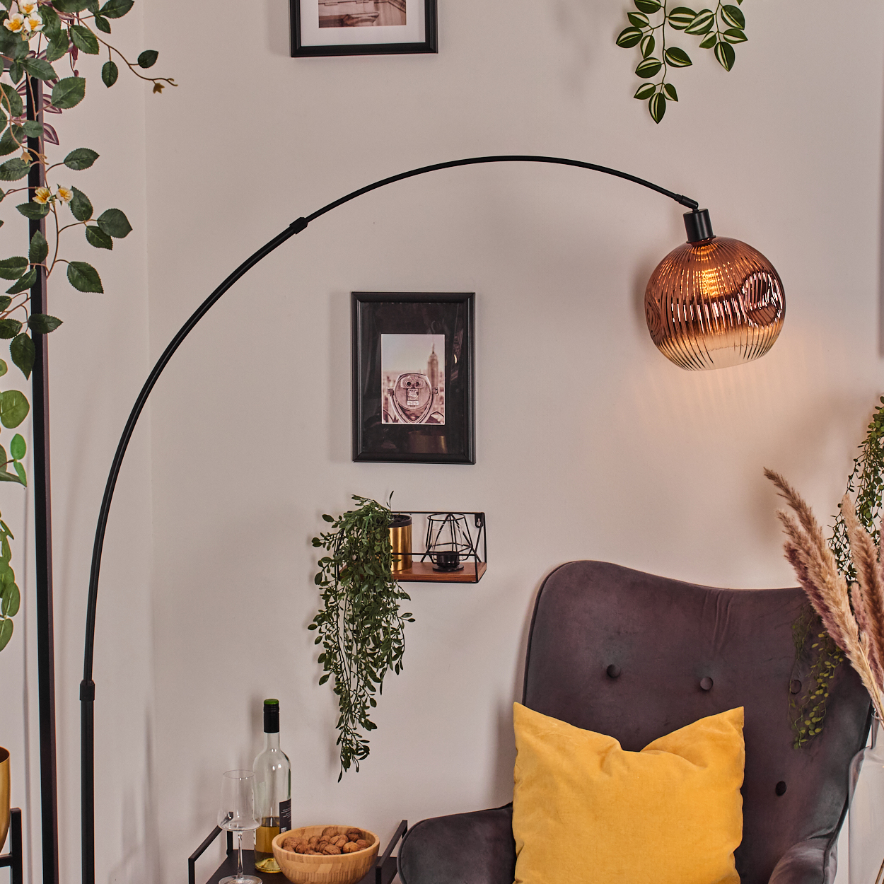 Ein Raum mit einer schwarzen Bogenlampe, einem grauen Sessel mit gelbem Kissen und einem kleinen Tisch mit einer Schale Nüsse.
