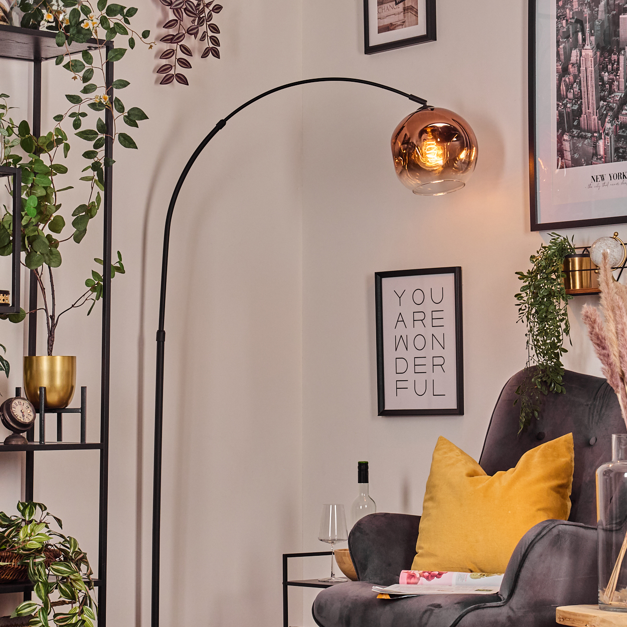 Ein modernes Wohnzimmer mit Stehlampe, Sessel und Regalen. Enthält Pflanzen, gerahmte Kunst und Dekorationsartikel. Die Lampe hat einen Kupferschirm.