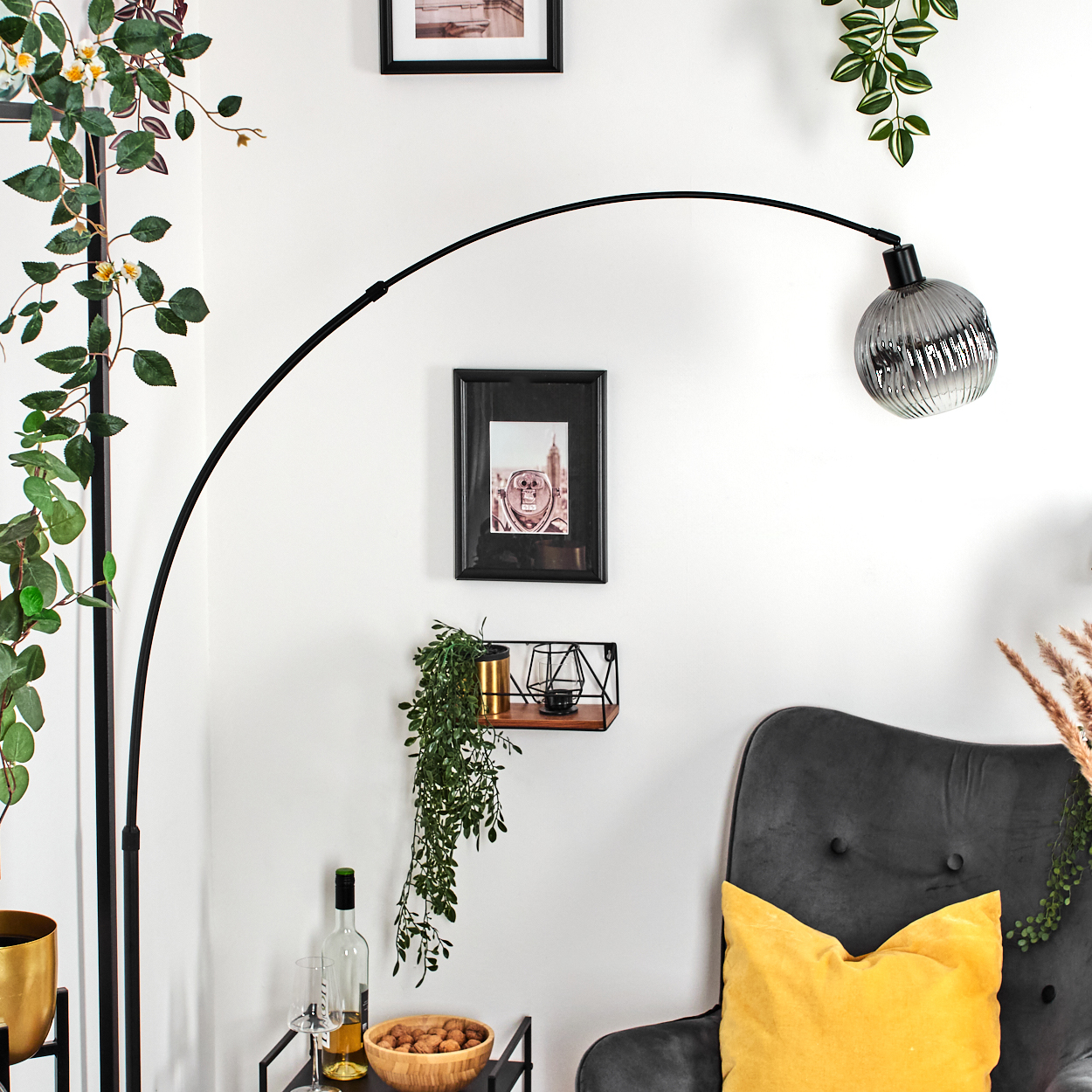 Ein Raum mit einer schwarzen Bogenlampe, gerahmten Bildern, einem grauen Sessel mit gelbem Kissen und Pflanzen an einer weißen Wand.