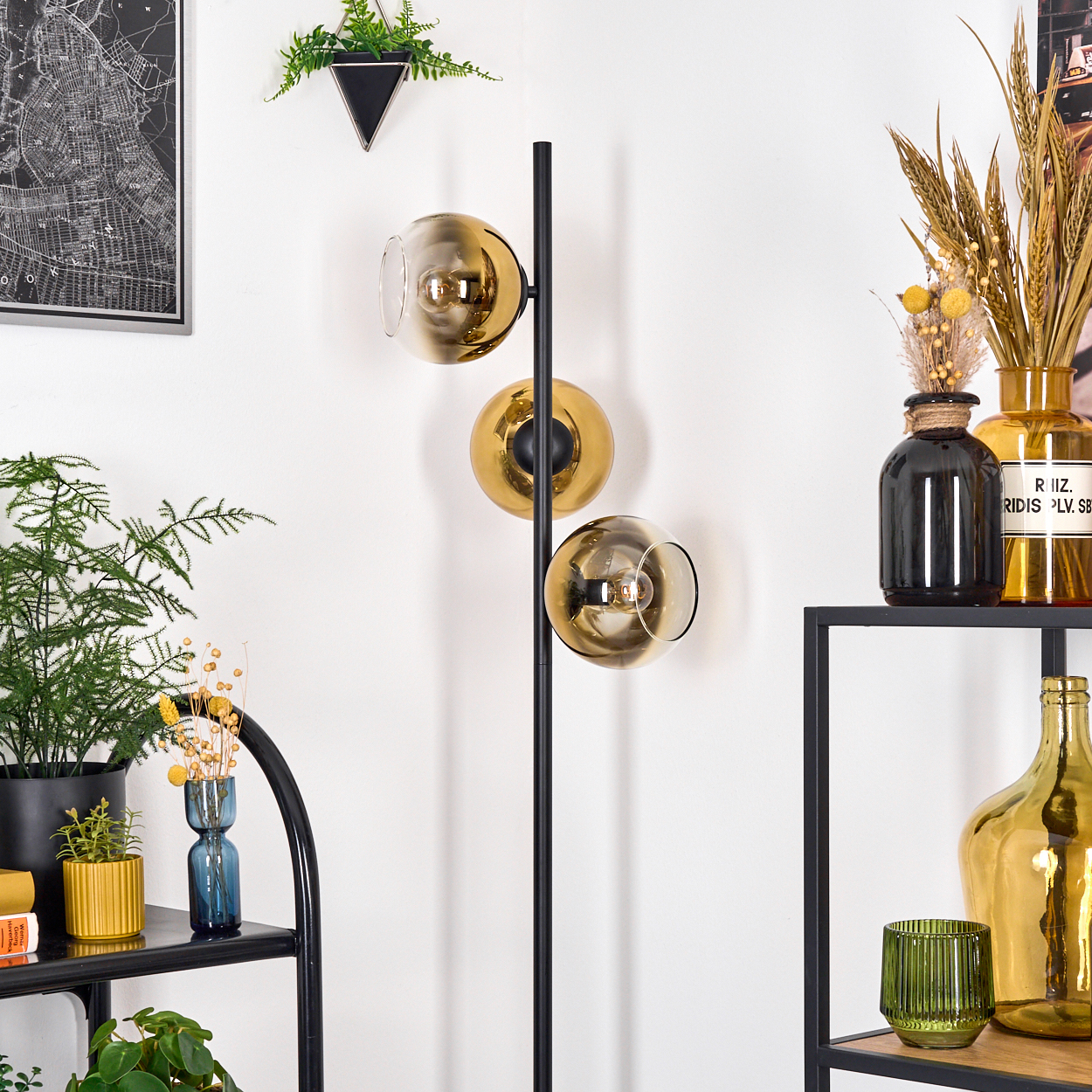 Ein modernes Zimmer mit einer schwarzen Metalllampe mit drei goldenen Glaskugeln. Regale halten Pflanzen, Vasen und Dekorationsartikel.