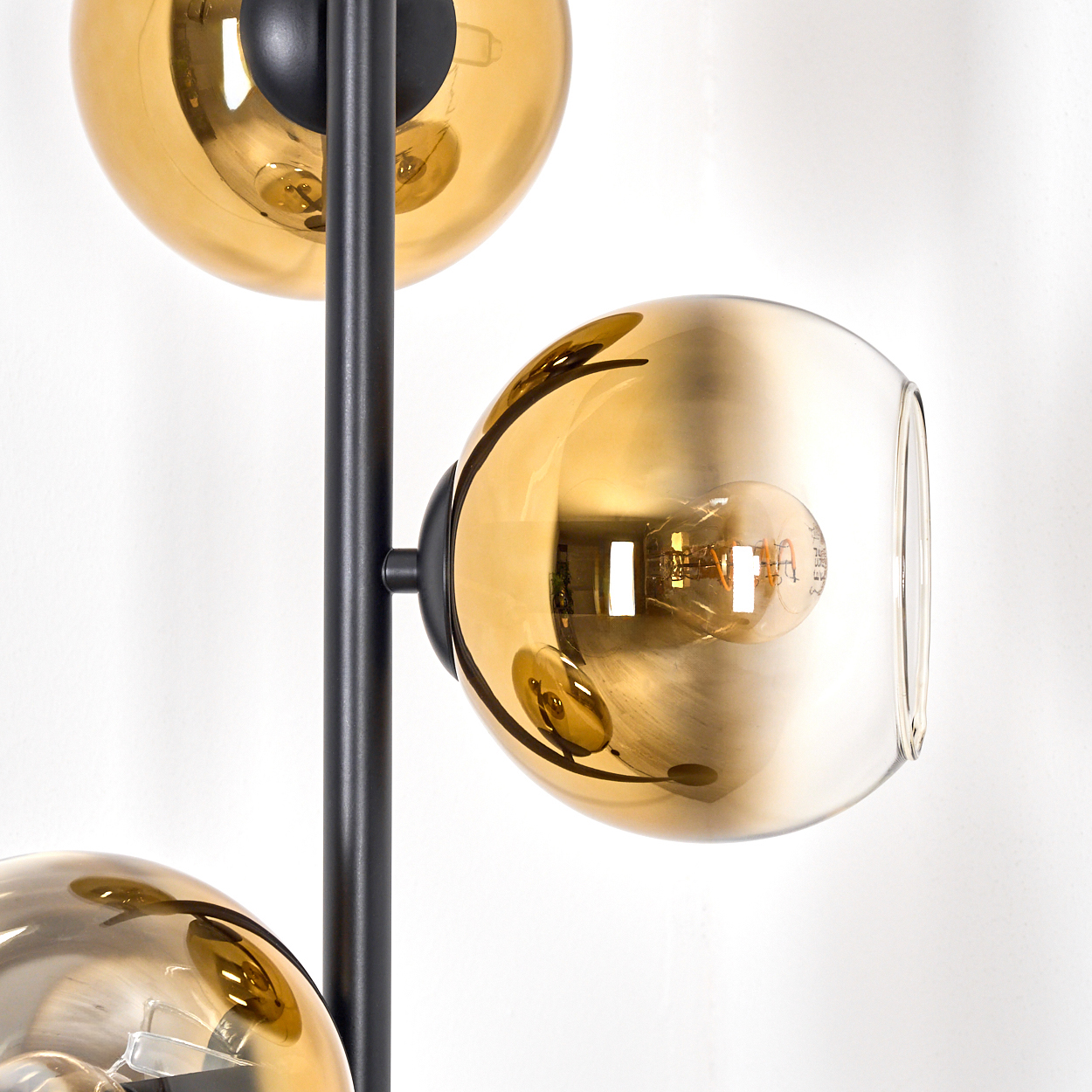 Eine moderne Lampe mit drei goldenen Glaskugeln und einem schwarzen Metallrahmen. Glühbirnen sind in den Kugeln sichtbar. Weiße Wand im Hintergrund.