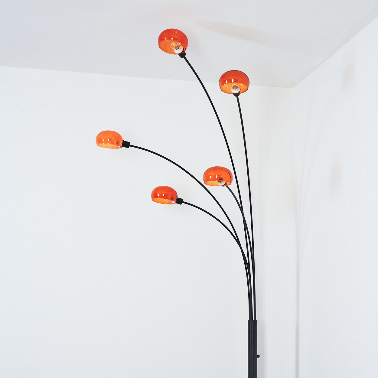 Eine moderne Stehlampe mit fünf orangefarbenen Glasschirmen. Der schwarze Rahmen ist nach oben gebogen. Der Hintergrund ist eine weiße Wand.
