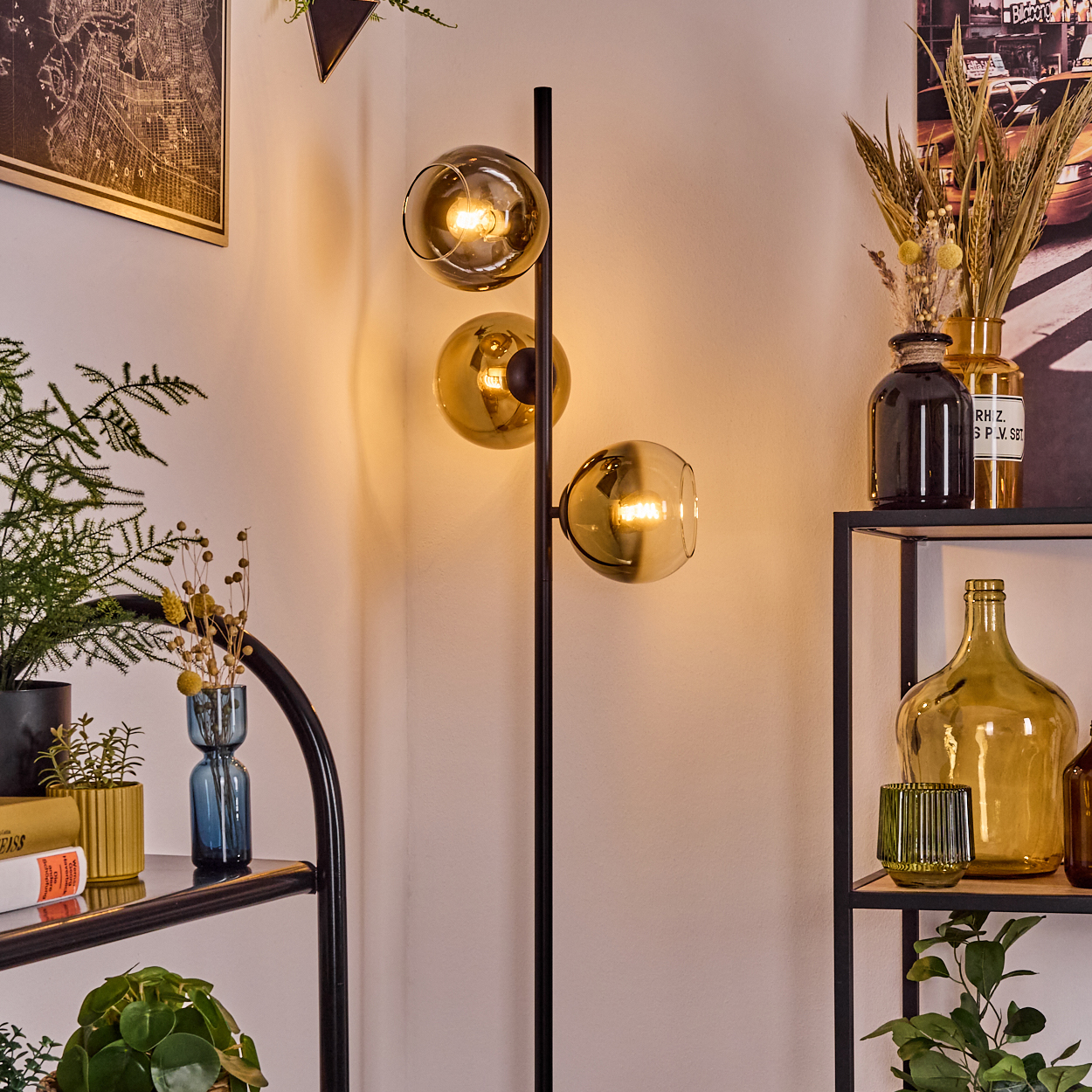 Eine moderne Stehlampe mit drei Kugelleuchten steht neben Regalen mit Pflanzen, Vasen und Dekorationen. Der Hintergrund ist eine weiße Wand.