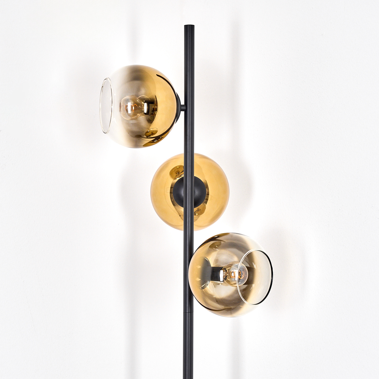 Eine moderne Stehlampe mit drei kugelförmigen, bernsteinfarbenen Glasschirmen, einem schwarzen Pfosten und einem weißen Wandhintergrund.
