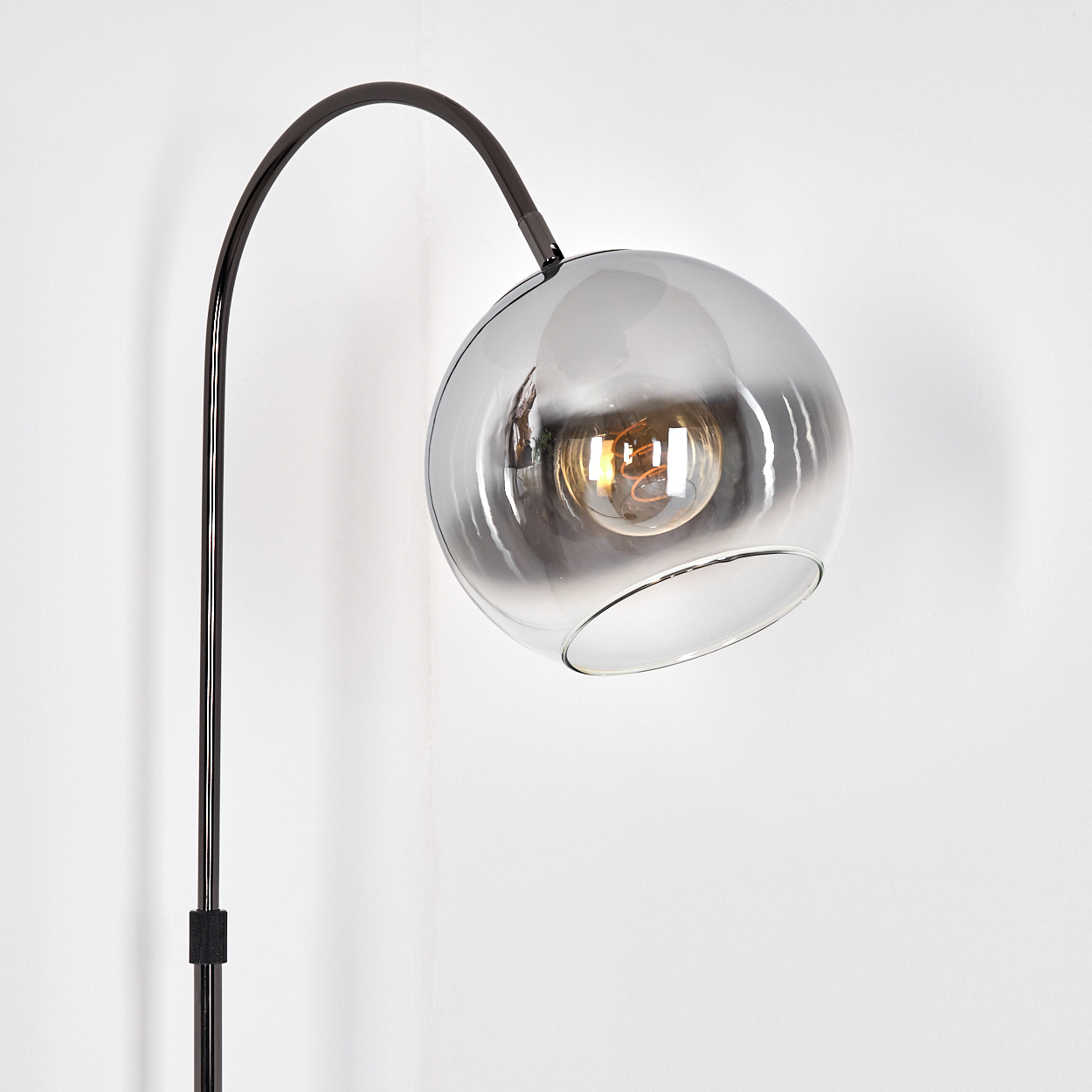 Eine moderne Stehlampe mit dunklem Metallrahmen und rundem, rauchfarbenem Glasschirm. Eine Glühbirne leuchtet im Schirm vor einer weißen Wand.