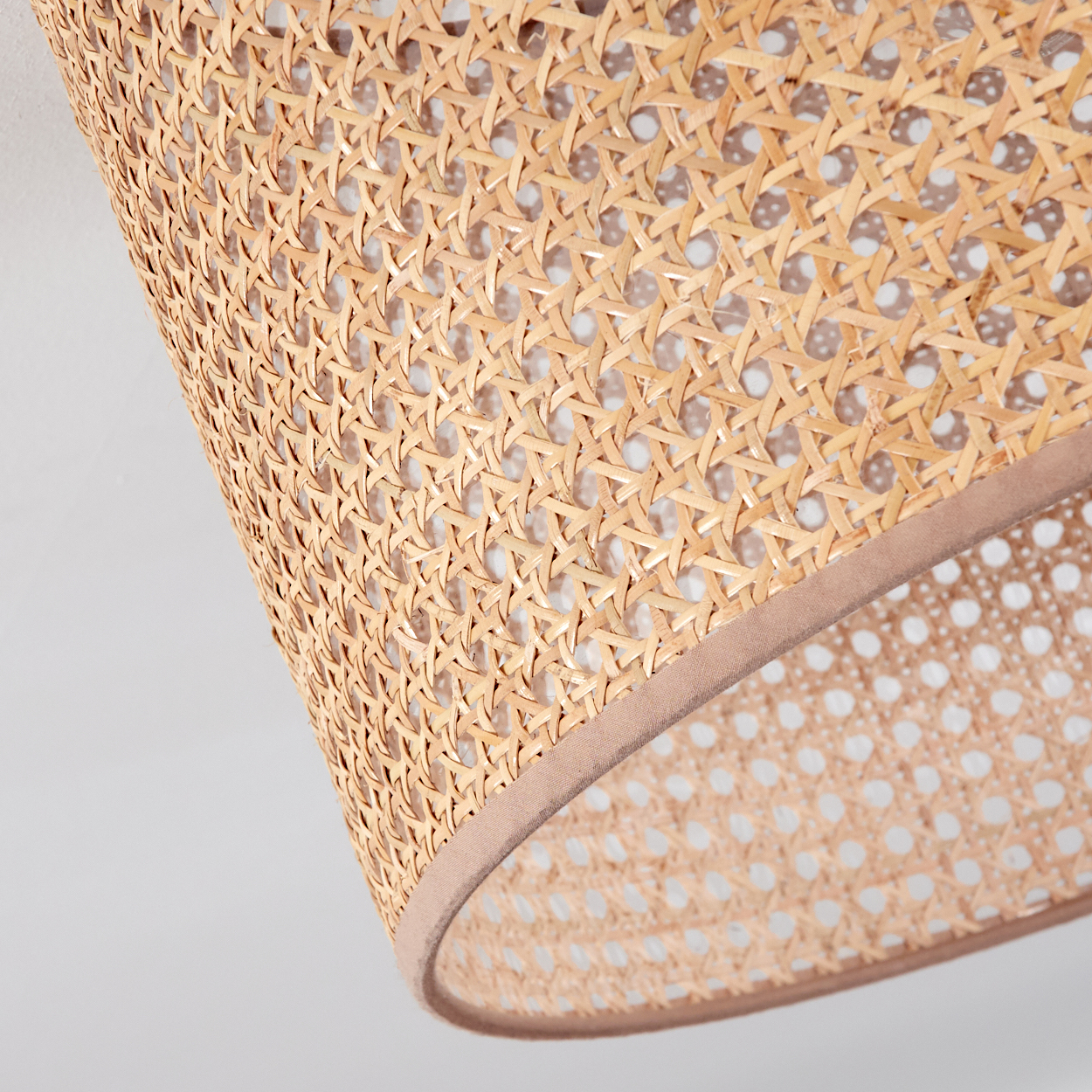 Nahaufnahme eines gewebten Rattan-Lampenschirms, tanfarben, mit hellbraunem Besatz. Der Hintergrund ist eine weiße Oberfläche.