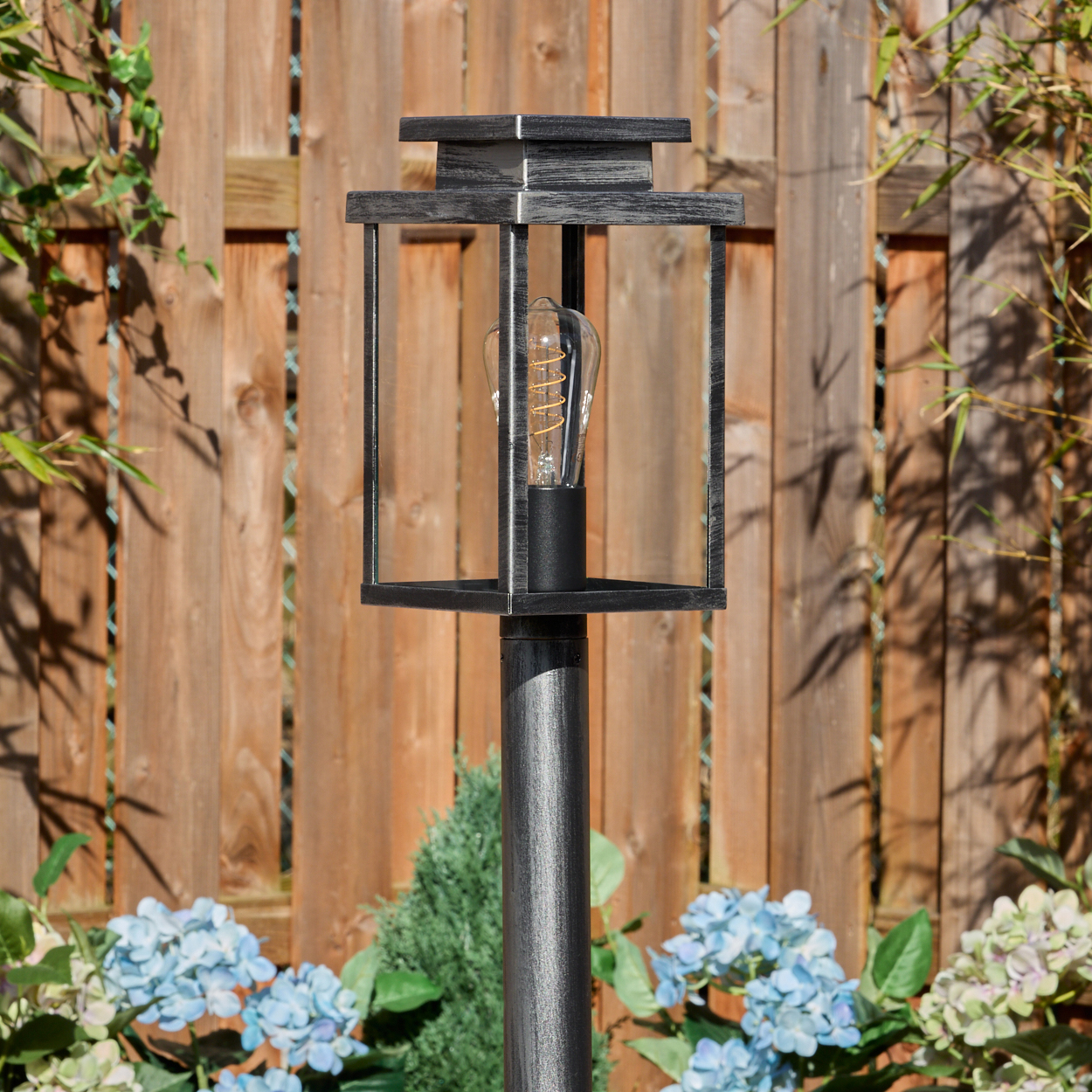 Eine dekorative Außenlampe mit Glühbirne steht vor einem Holzzaun, umgeben von Pflanzen und blauen Blumen.