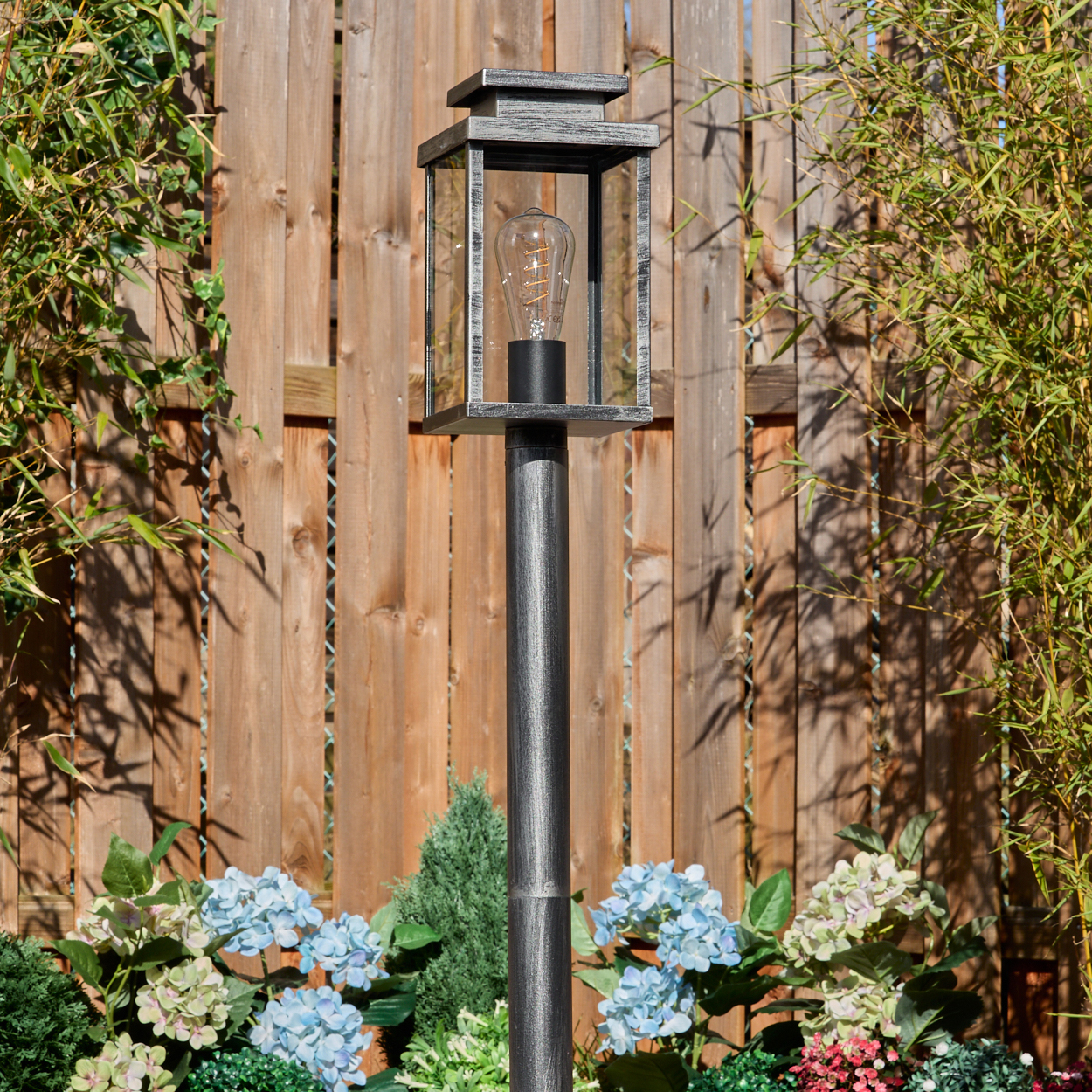 Außenlampe mit Glasgehäuse und Glühbirne. Holzzaun im Hintergrund. Blumen und Grün umgeben die Lampe.