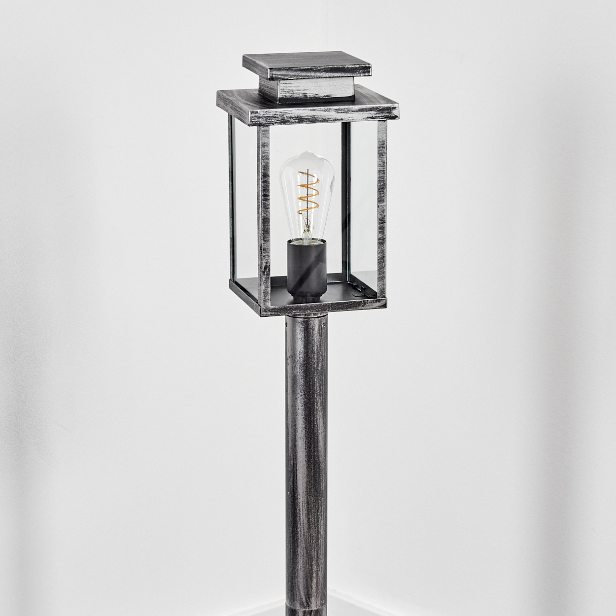 Eine metallisch graue Außenlampe mit einer Glühbirne steht vor einer weißen Wand. Die Lampe hat ein rechteckiges Design und einen langen Pfahl.