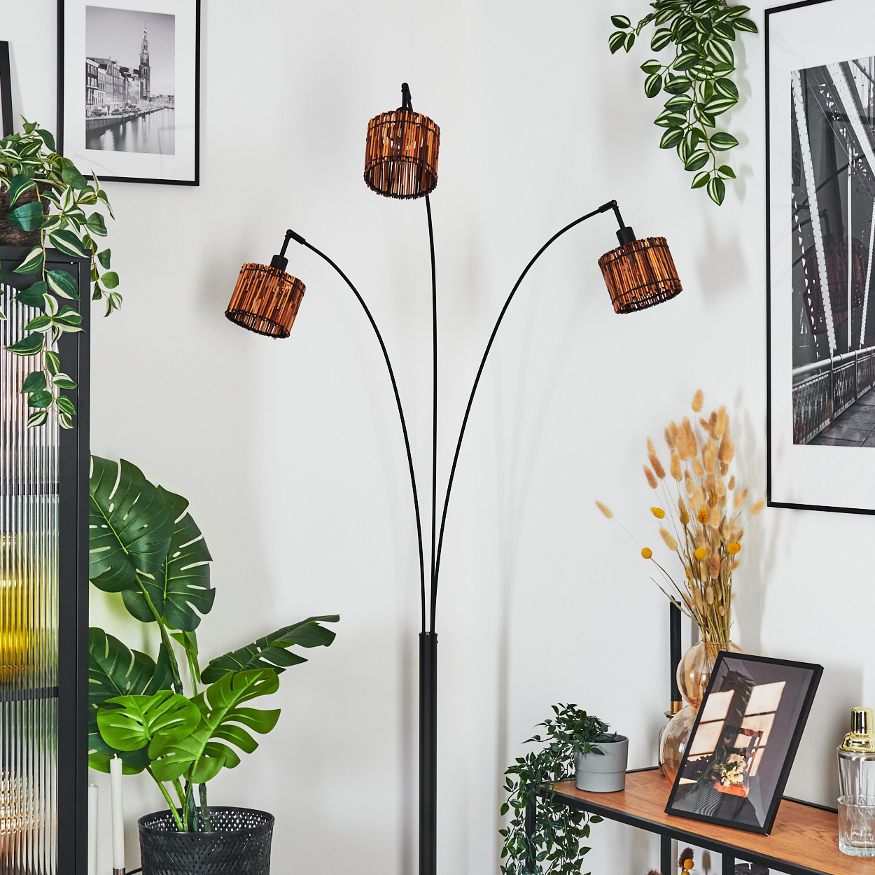 Ein modernes Wohnzimmer mit einer schwarzen Stehlampe mit drei Lampenschirmen. Pflanzen und gerahmte Bilder schmücken die weißen Wände.