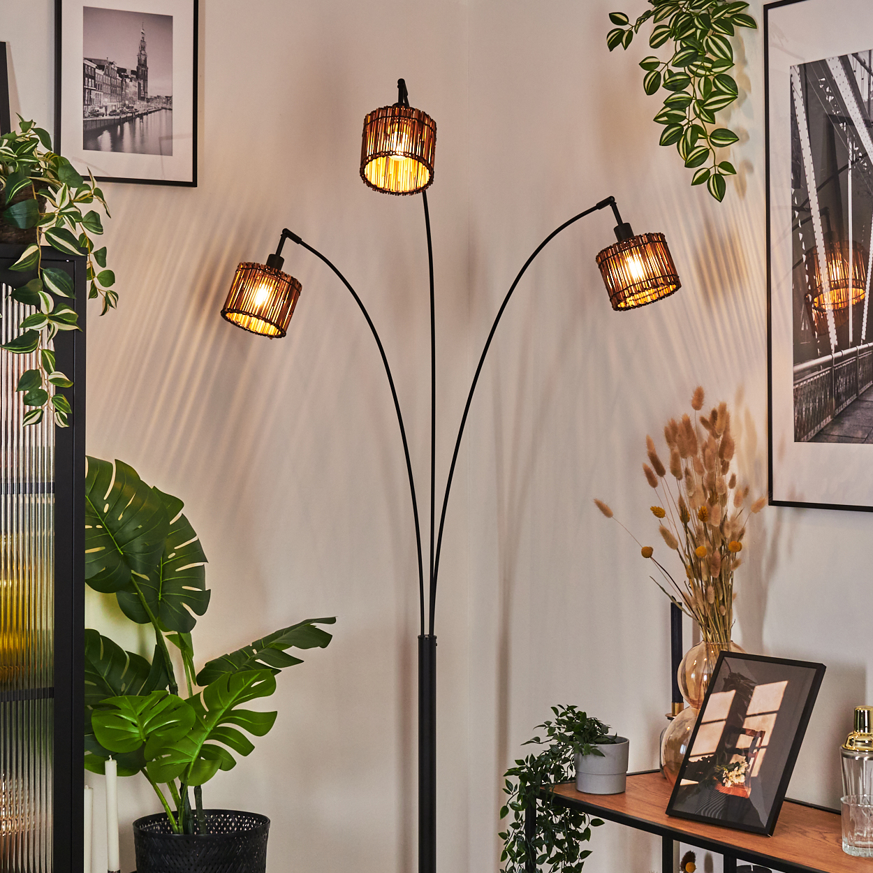 Eine Stehlampe mit drei Lampenschirmen befindet sich in einem Raum mit Pflanzen und Bildern. Die Lampenschirme sind braun, die Wände weiß.